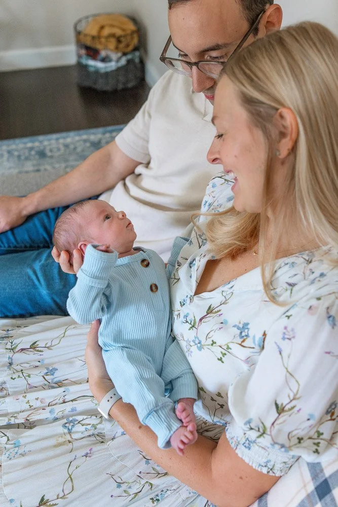 A woman holding a newborn baby boy with a man sitting nearby looking fondly at them, in a cozy room.