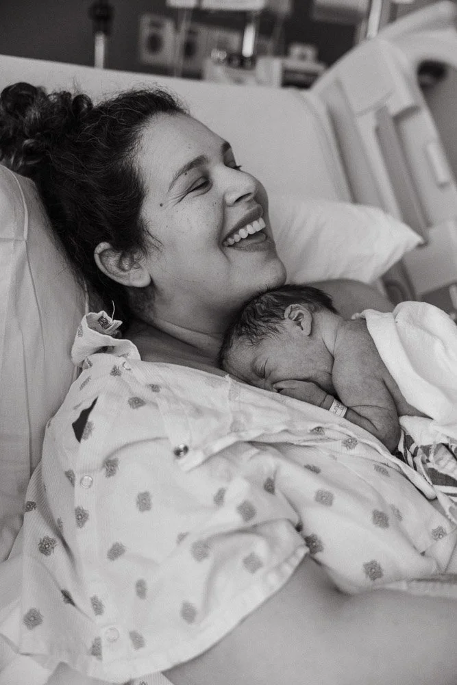 A woman in a hospital bed, smiling with her eyes closed, holding a newborn baby against her chest. The woman appears to be a new mother in a hospital setting.