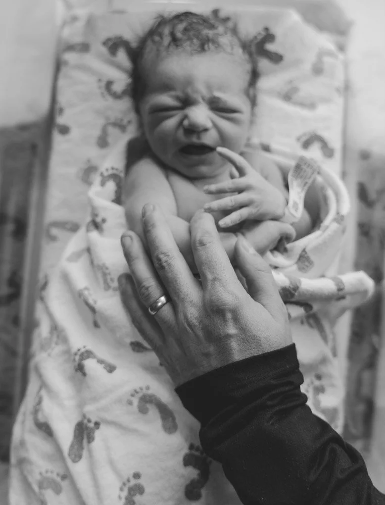 A newborn baby crying while being gently held by an adult hand, wrapped in a patterned blanket, on a bed or changing table.