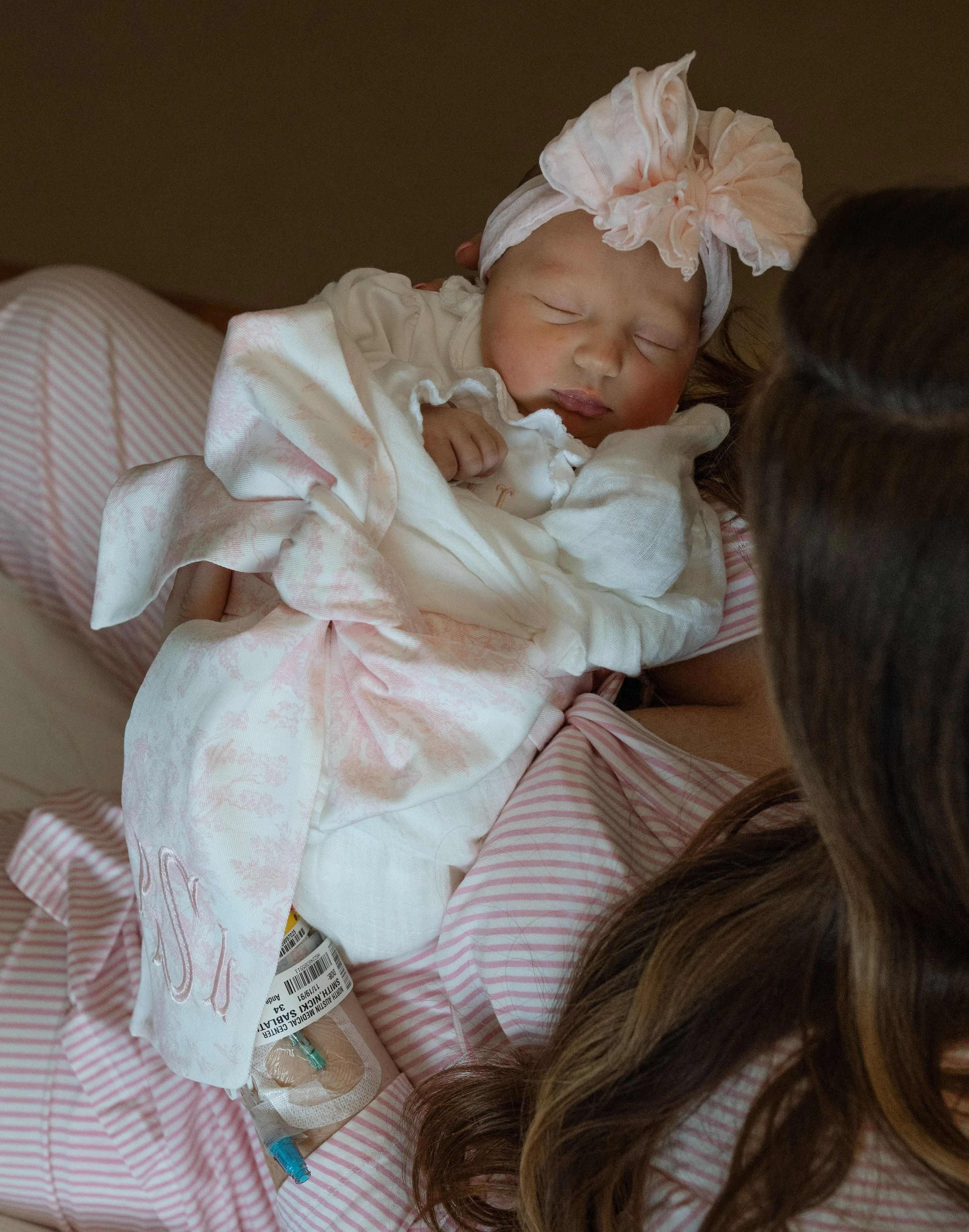 a newborn baby girl wearing a pink bow headband and monogrammed bow swaddle is held by a woman wearing matching pink pjs / pajamas and matching robe 