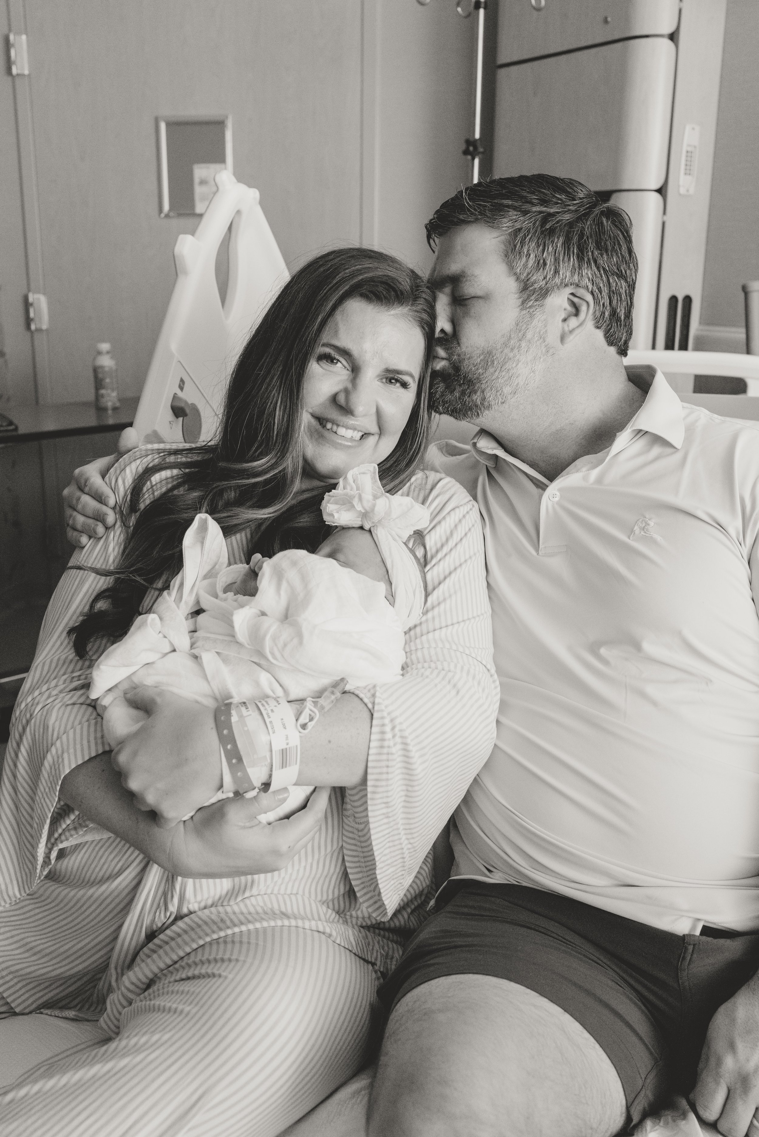 in hospital fresh 48 session where new parents sit on a hospital bed, woman is holding a baby that is swaddled and the man is kissing the woman's head