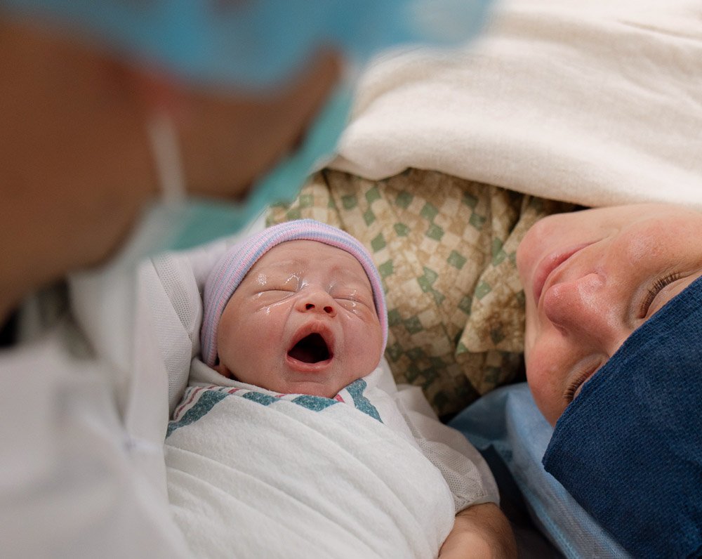 A newborn baby yawning while lying in a hospital bed, with a woman, likely the mother, resting her head nearby.