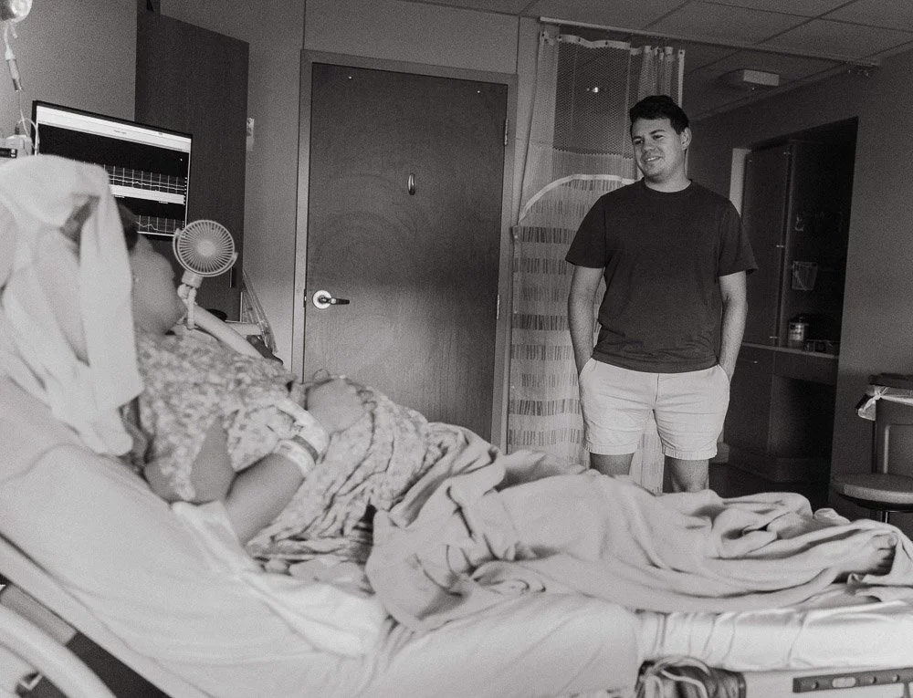A person lying in a hospital bed with medical tubes and monitors, while a young man in casual clothing stands nearby, smiling.