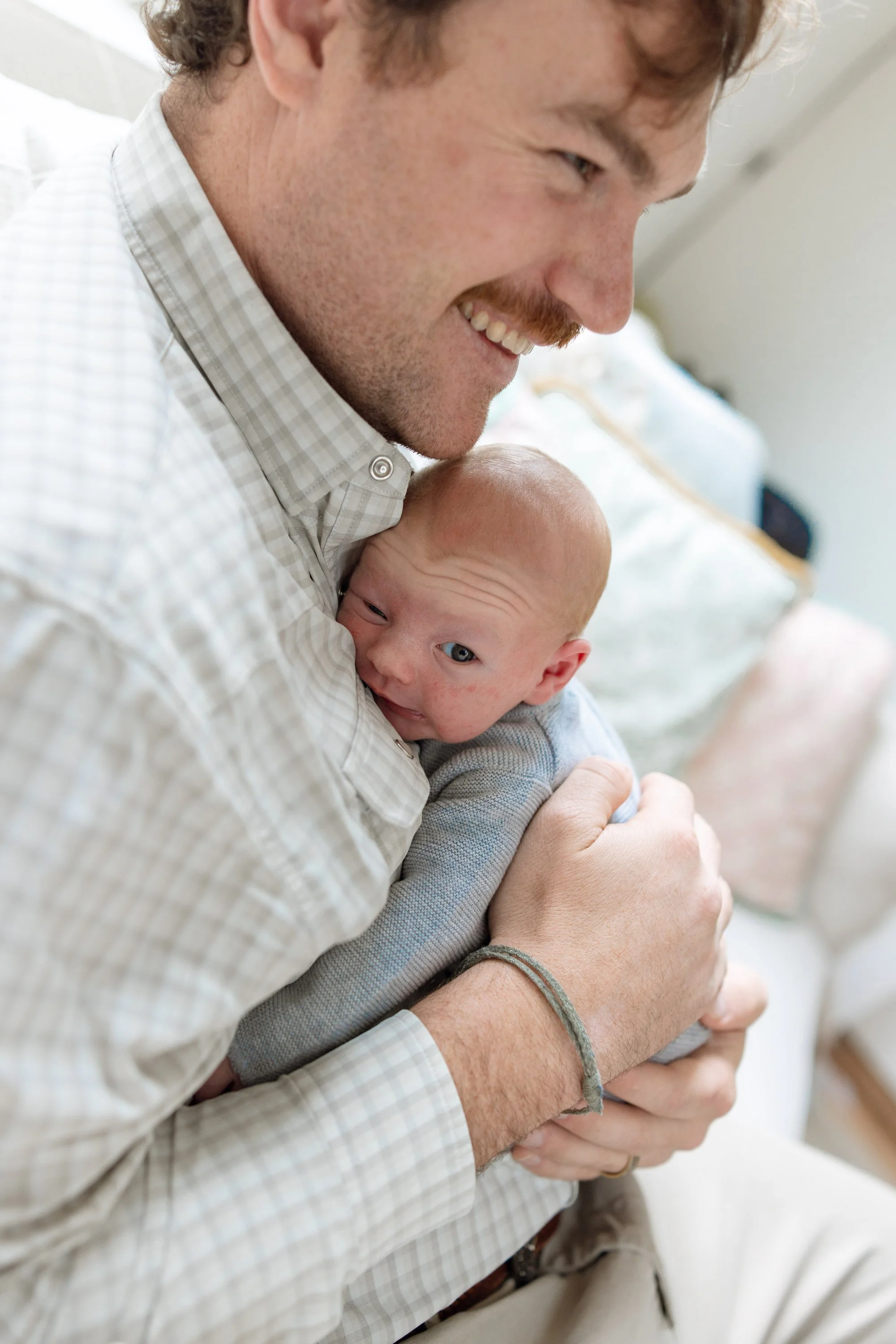 A man holding a newborn baby close, both smiling. The man is wearing a checkered shirt, and the baby is cuddled against his chest, looking towards the camera.