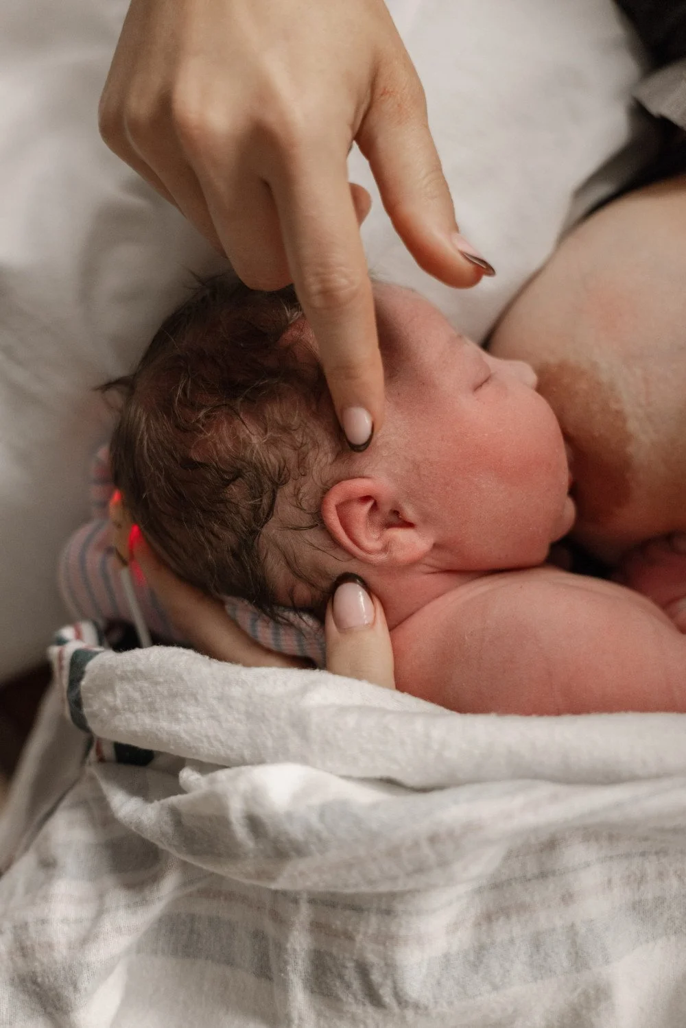 A newborn baby being held and gently touched on the head by an adult's hand in a hospital setting.