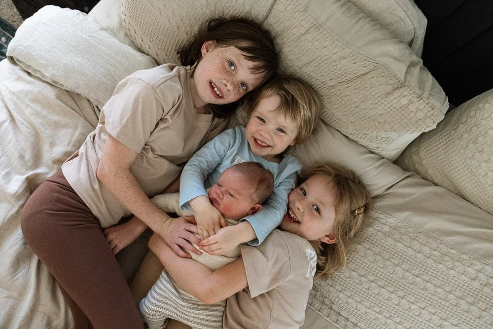 Four children lying on a bed, smiling and hugging each other, with a newborn baby.