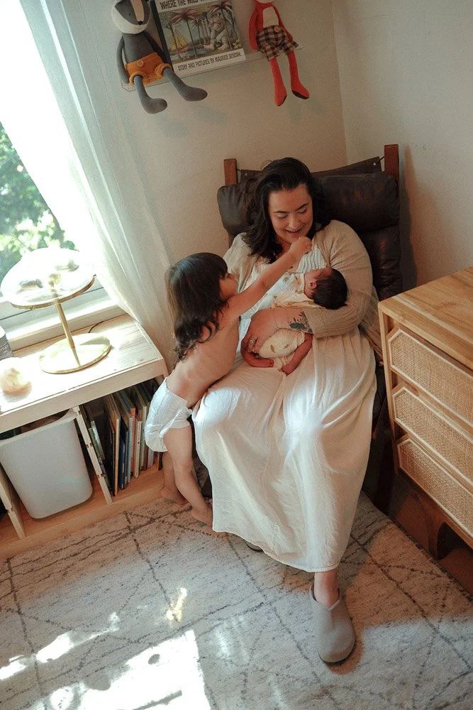 A woman sitting on a chair holding a newborn, a young child reaching out to touch the baby's face, in a cozy room with a window, bookshelf, and wall decorations.