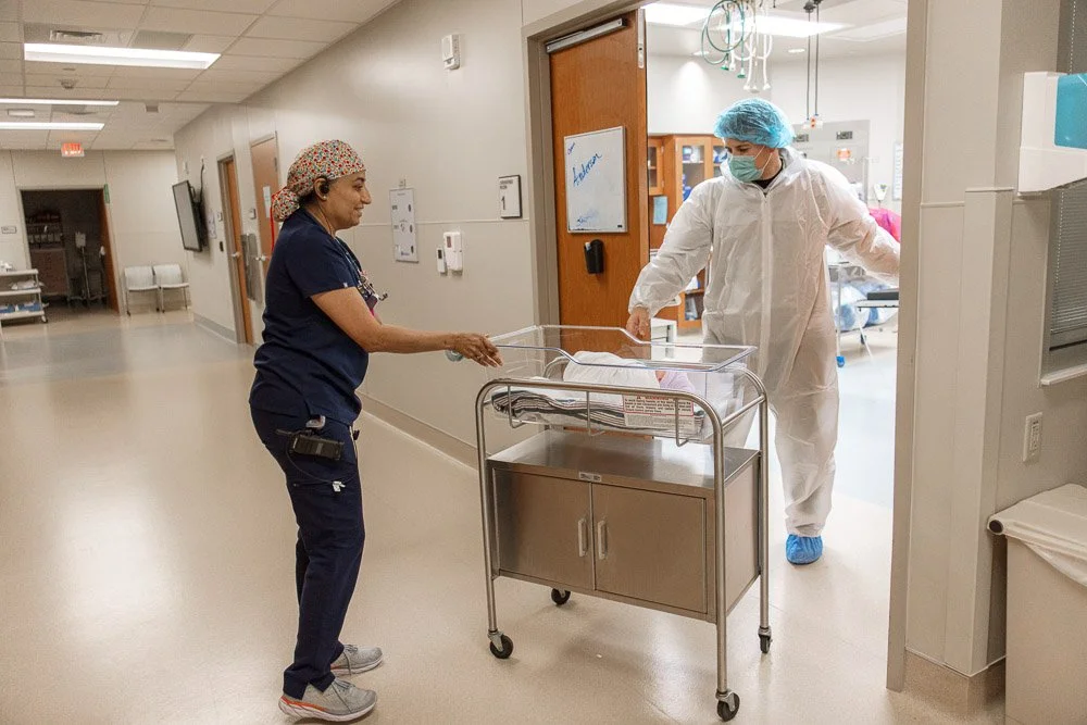 A healthcare worker and a nurse exchanging a newborn baby in a hospital corridor, with the nurse pushing a baby bassinet.