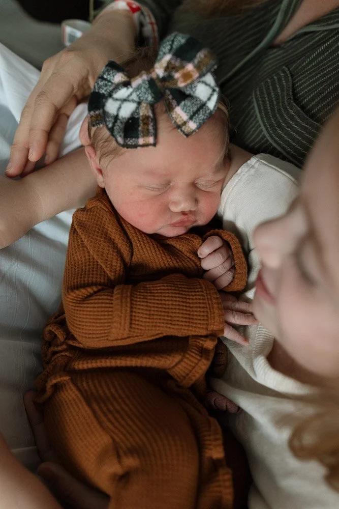 Close-up of a newborn baby sleeping peacefully, wearing a plaid bow headband and brown outfit, with an older child gently looking at the baby, in a cozy, intimate setting.