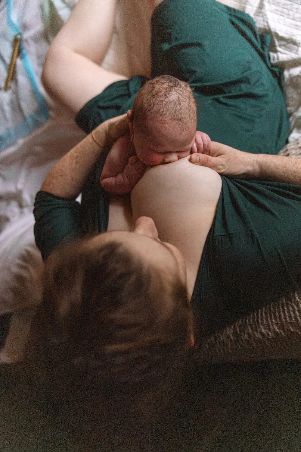 A person is laying on their side, breastfeeding a newborn baby who is sucking on their breast, with an older child lying nearby.