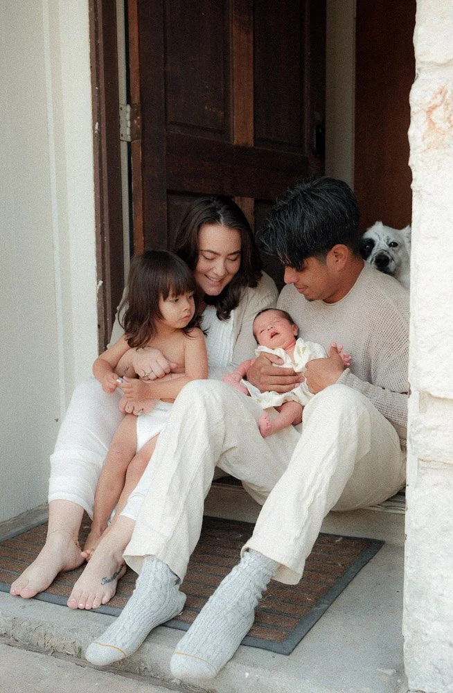 a woman, a young boy and a man holding a newborn baby are sitting one their front door stem all smiling at each other, the family dog stands behind them