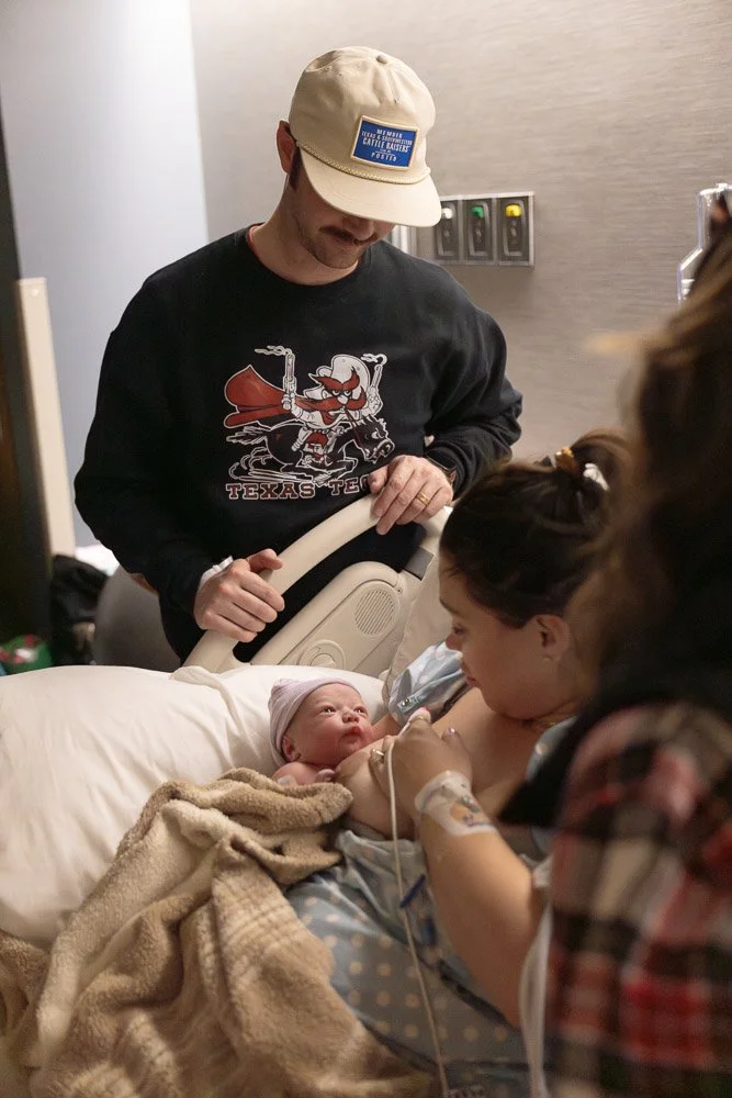 A woman in a hospital bed holding an infant with medical monitors attached, looking at the baby. A man wearing a beige hat and a Texas Tech sweatshirt stands nearby, watching the woman and baby. The scene appears to be in a hospital room.