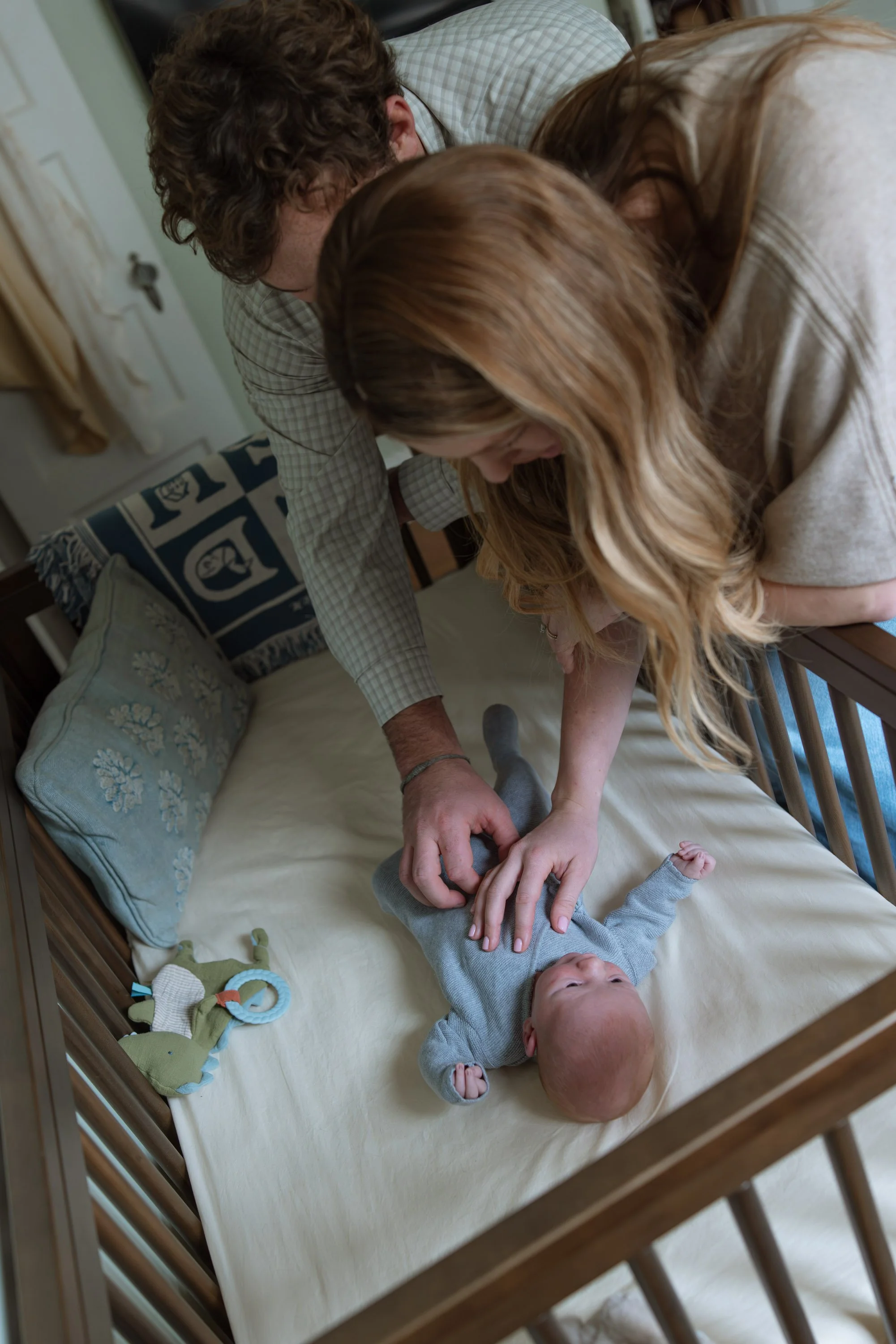 mom and dad lean over a baby crib looking at their newborn son in a blue knit outfit. 