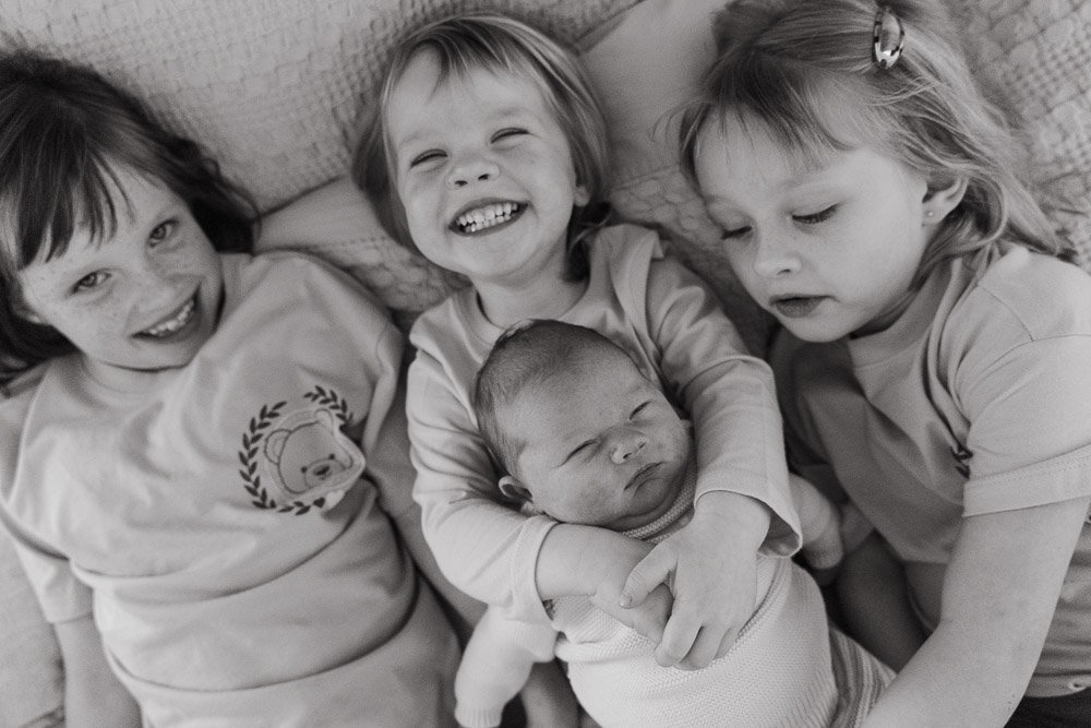 Four young children, two girls and a boy, lying down together on a bed with a newborn baby in the middle. The children are smiling and appear happy.