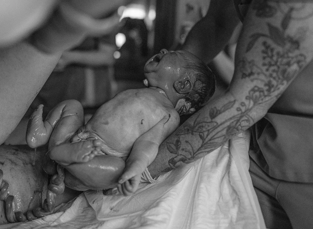 A newborn baby just delivered, being held by a person with arm tattoos, in a birth center setting.
