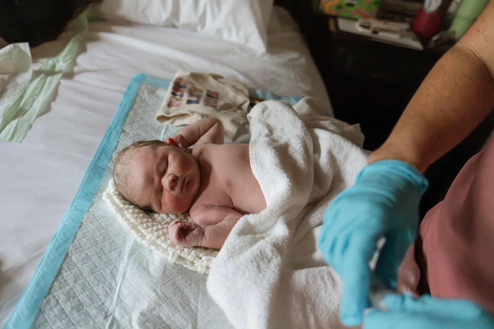 Newborn baby lying on a hospital bed, wrapped in a white towel, with a brownish blanket nearby, being gently held by a person wearing blue gloves.