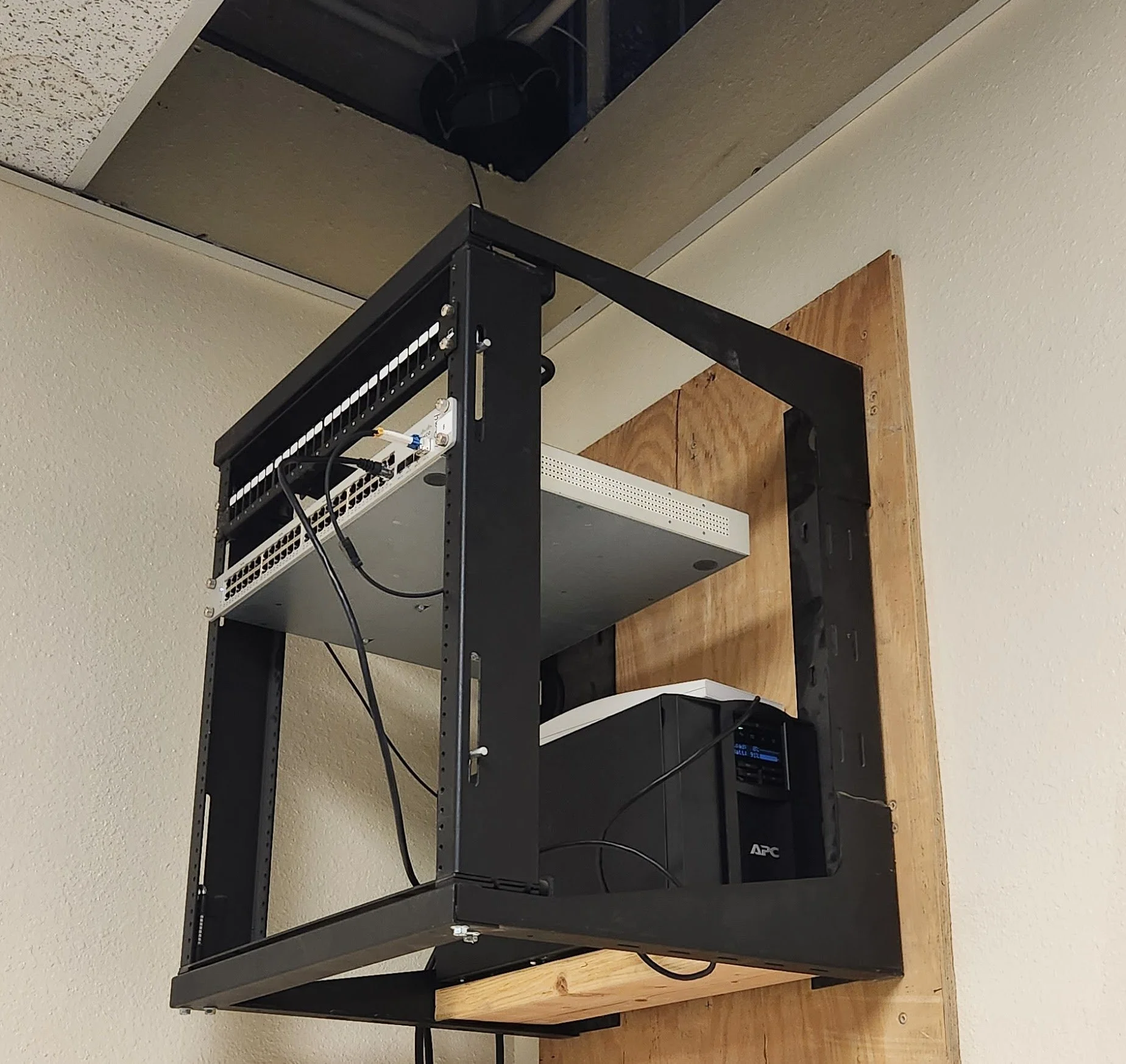 A black metal open-frame rack holding network equipment, including a switch and a power distribution unit, mounted on a wooden backboard on a wall.