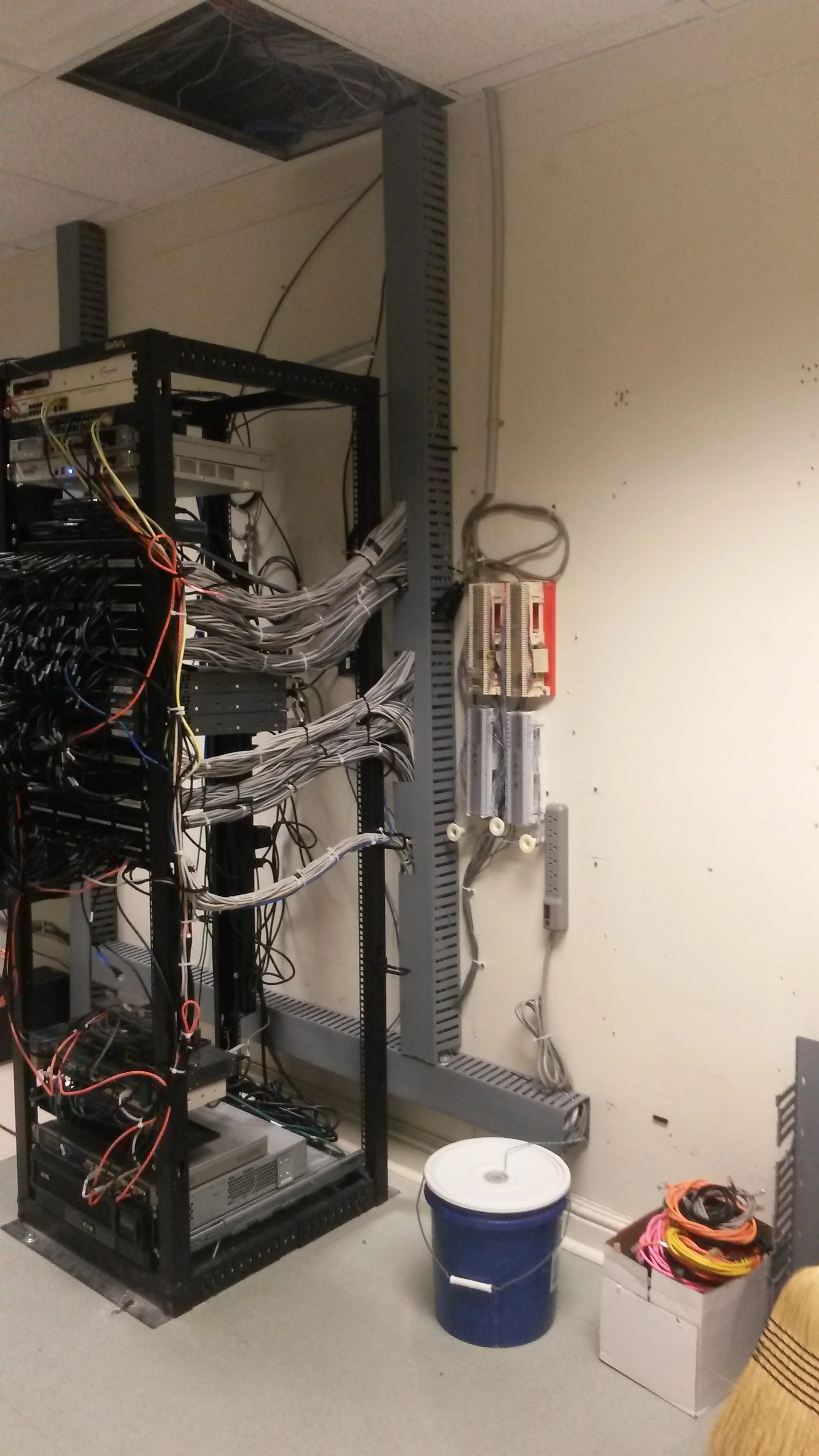 A server rack with tangled cables, an electrical panel on the wall, a bucket on the floor, and various wires on a box to the right.