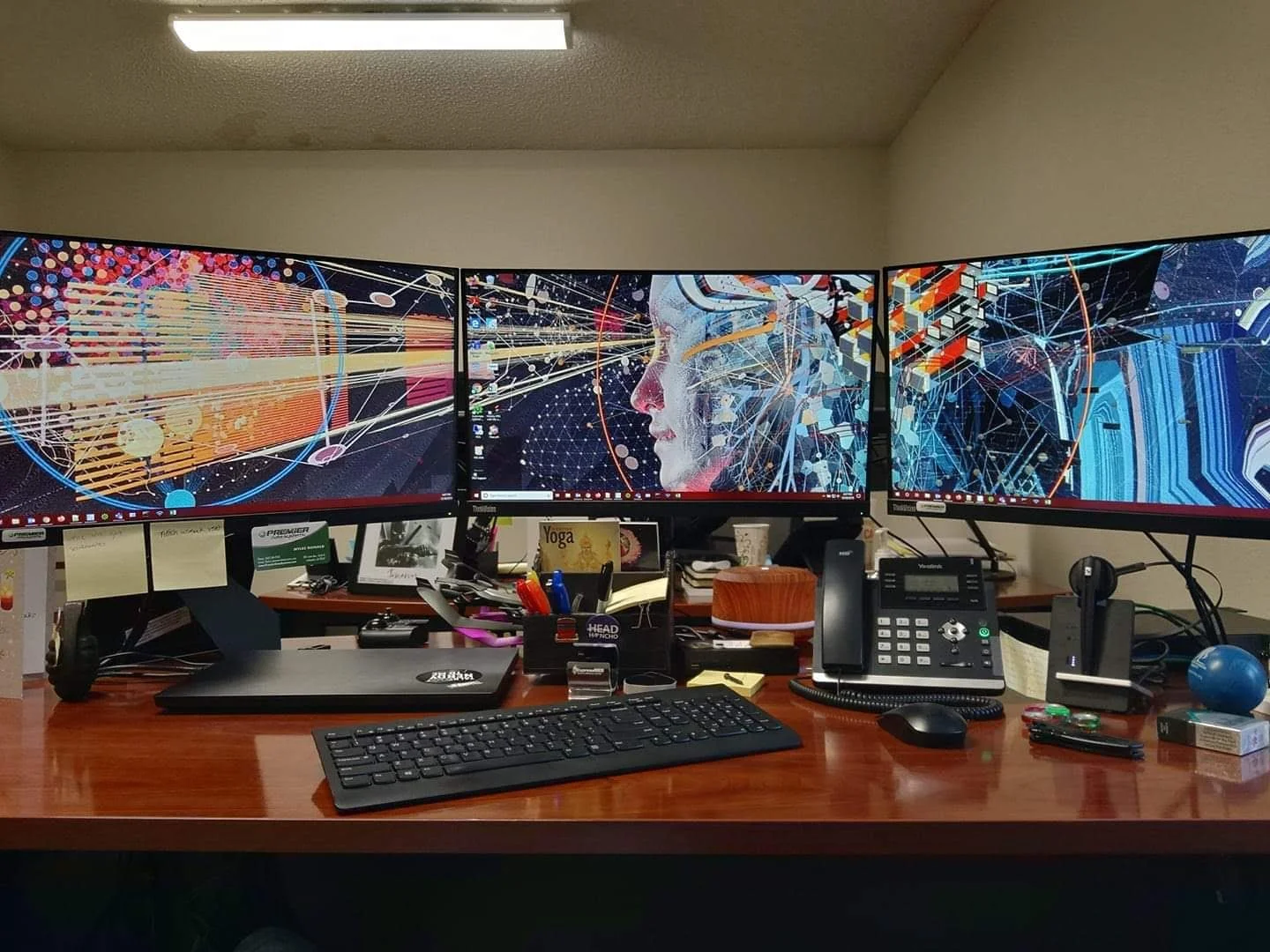 Office workspace with a large triple monitor display showing digital artwork of a human face with abstract circuit and network design. The desk has a keyboard, mouse, office phone, and various office supplies.