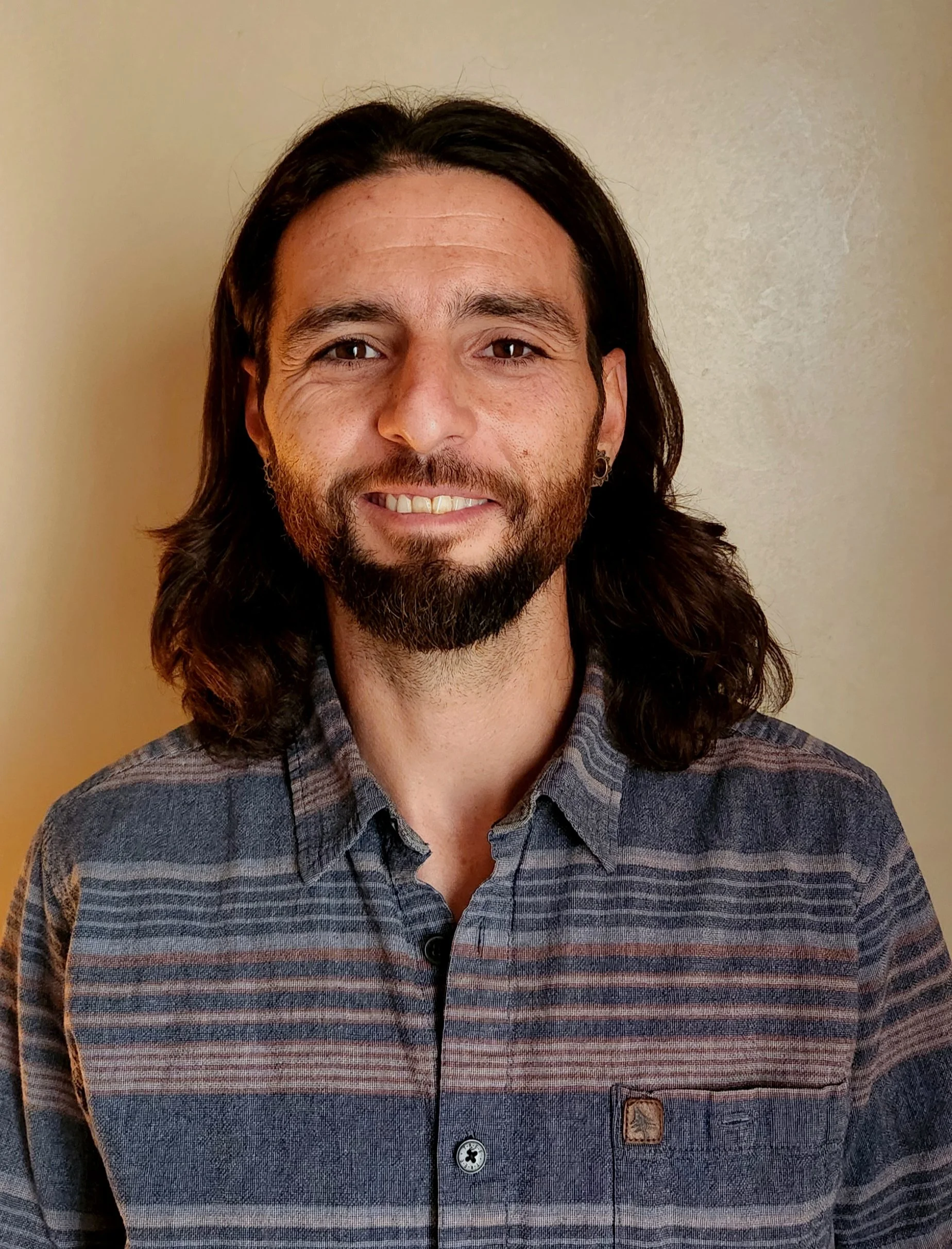 Man with long dark hair, beard, and mustache smiling, wearing a striped button-up shirt, standing against a plain wall.