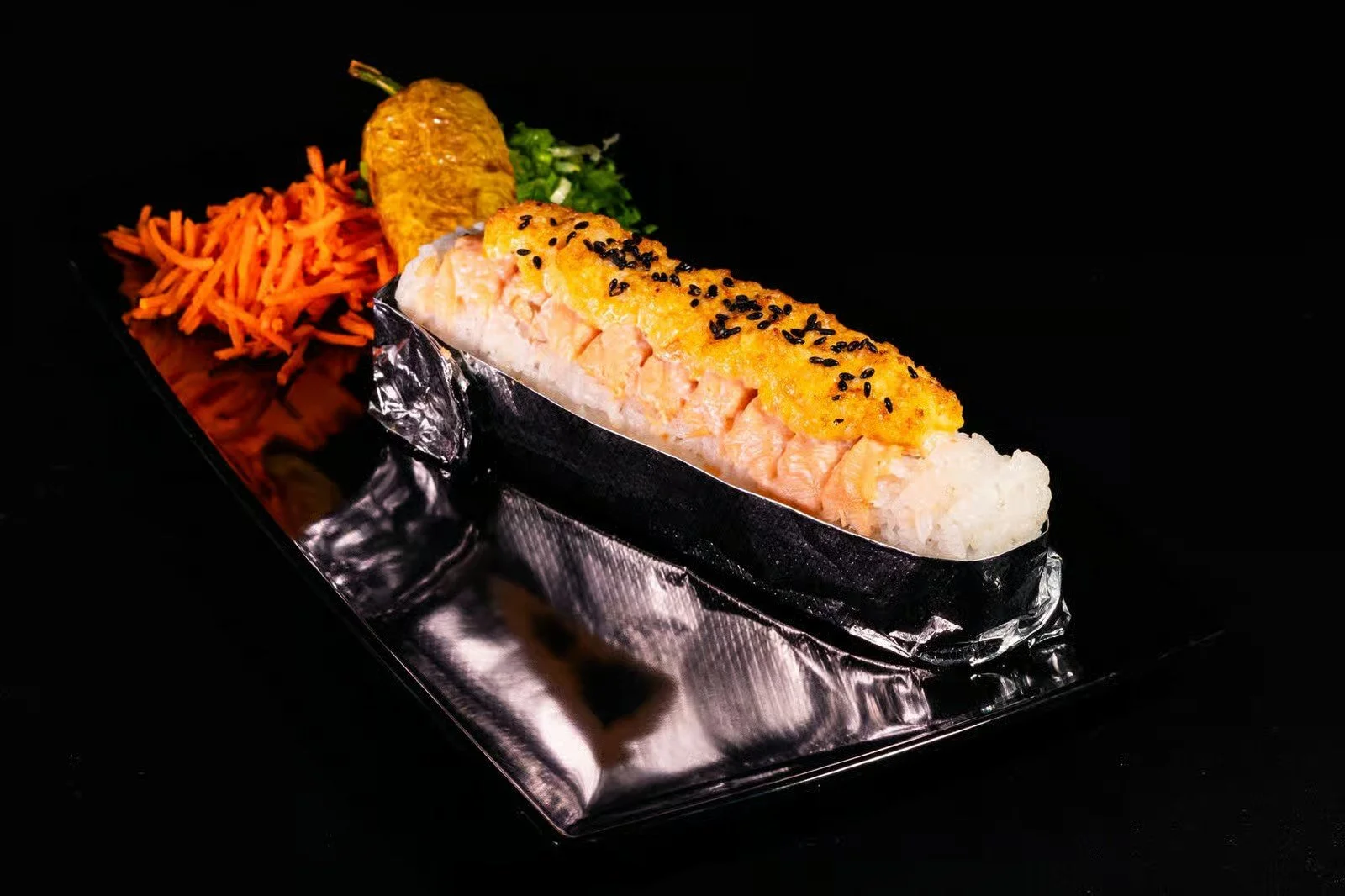 Salmon sushi roll topped with sesame seeds, served with shredded carrots, a fried pepper, and chopped green onions on a black background.