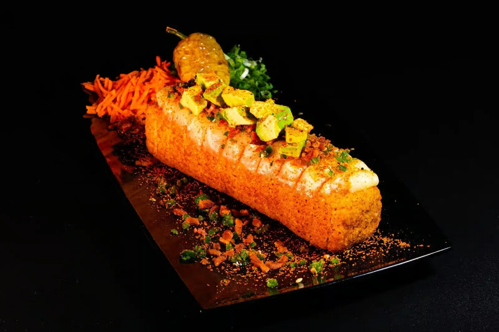 Crispy fish fillet topped with chopped avocados, served with shredded carrots, a lemon wedge, and garnished with herbs on a black plate.