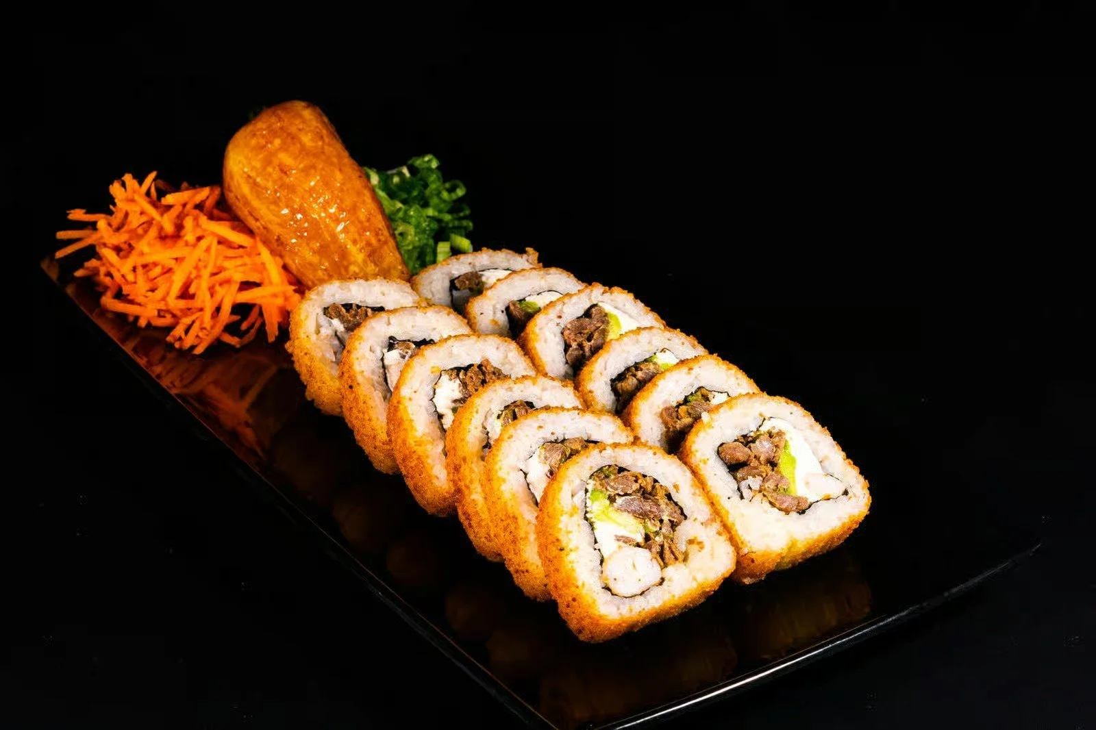 Sushi rolls with fried coating, sliced to show filling of beef, rice, and vegetables, on a black rectangular plate with shredded carrots, a sweet potato, and chopped green onions.