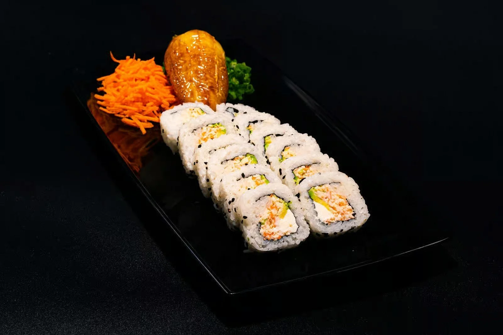Sushi rolls with vegetables and seafood on a black plate, garnished with shredded carrots, a banana, and green lettuce on a dark background.