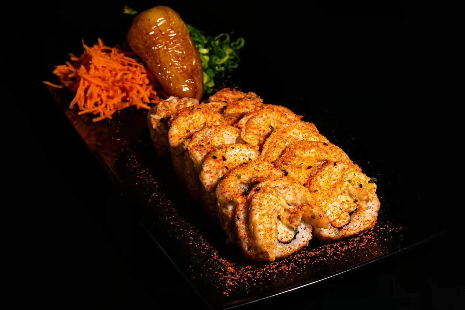 Sushi roll with spicy topping, a jalapeño pepper, shredded carrots, and chopped green onions on a black plate.