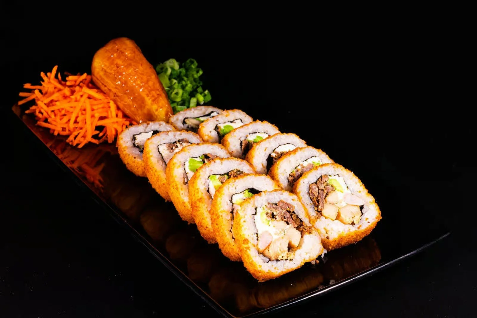 Sushi rolls with vegetables and seafood on a black rectangular plate, accompanied by shredded carrots, chopped green onions, and a fried plantain.