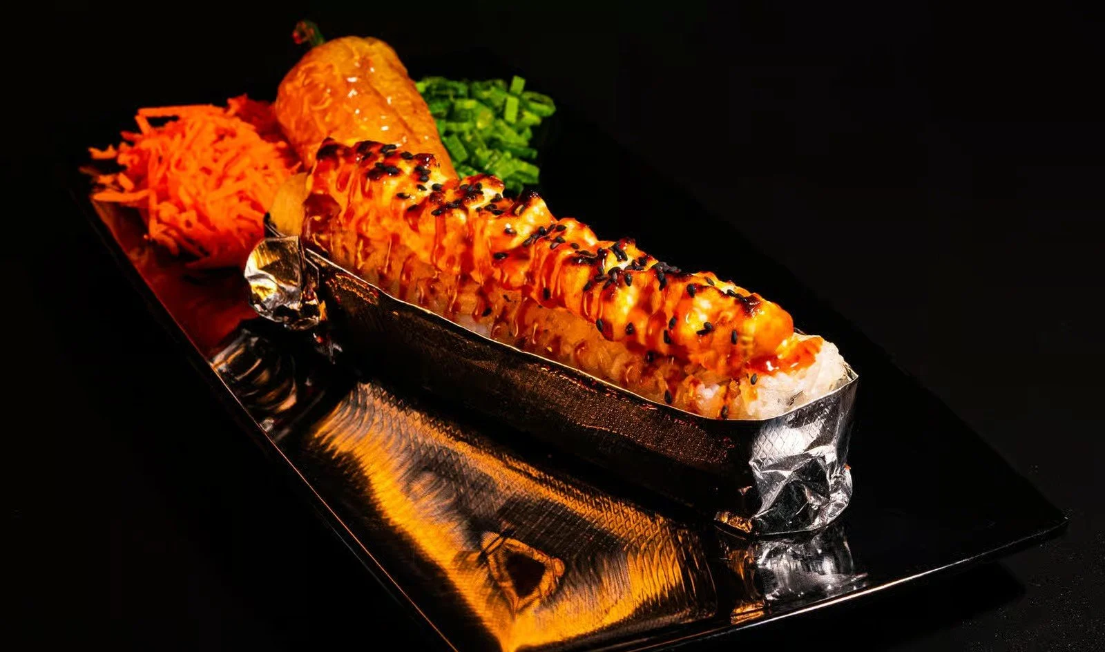 Sushi roll topped with sauce and black sesame seeds, accompanied by shredded carrots, chopped green onions, and pickled ginger on a black background.