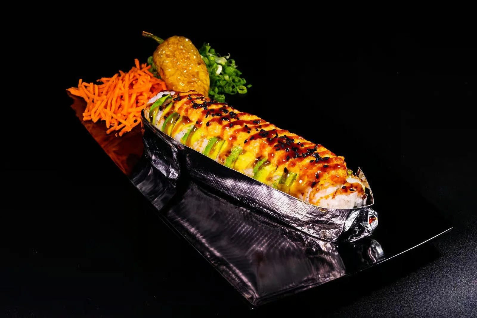 Sushi roll with toppings, shredded carrots, green onions, and a pickled yellow chili pepper on a black background.