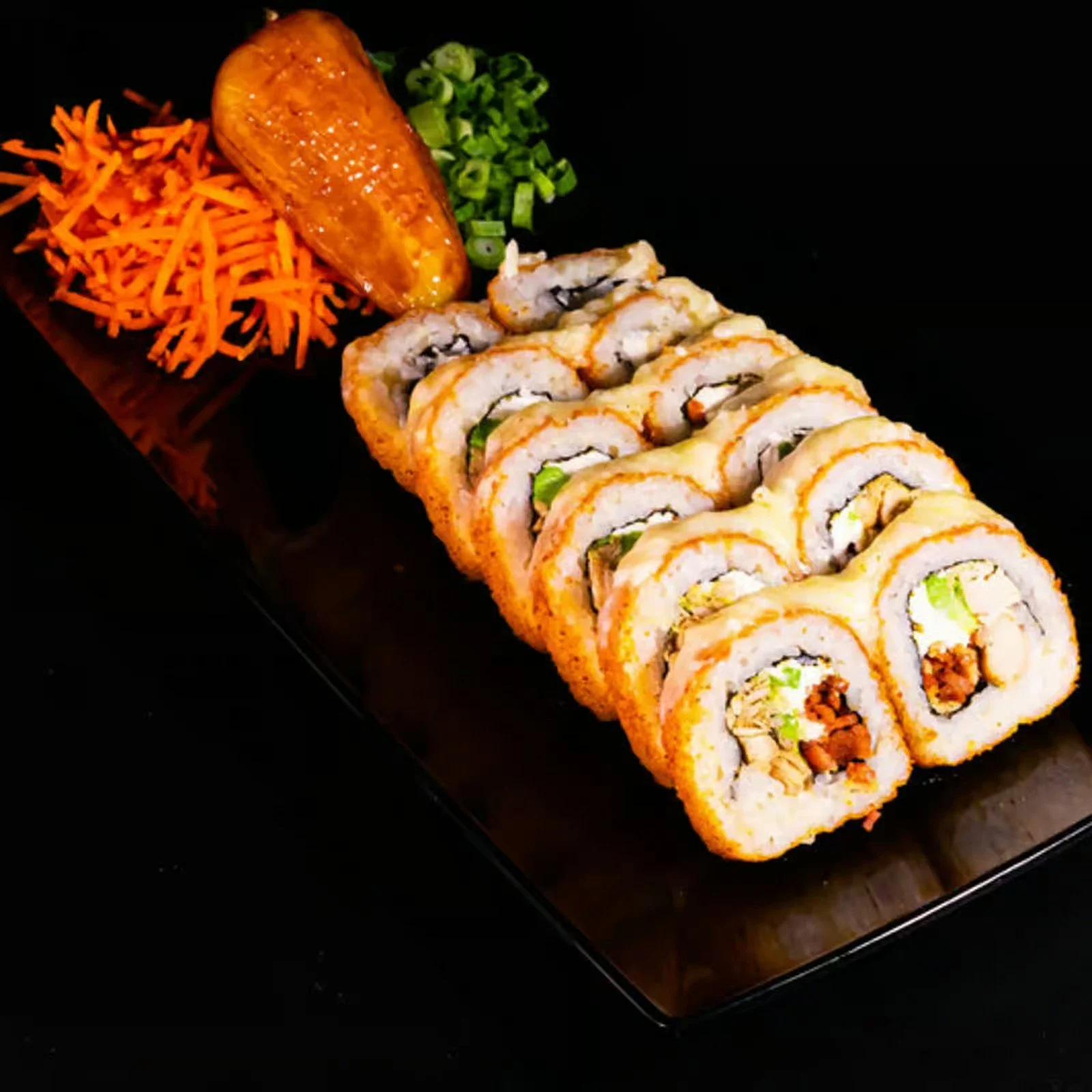 Sushi rolls on a black plate with shredded carrots, a sweet potato, chopped green onions, and a piece of fried plantain.