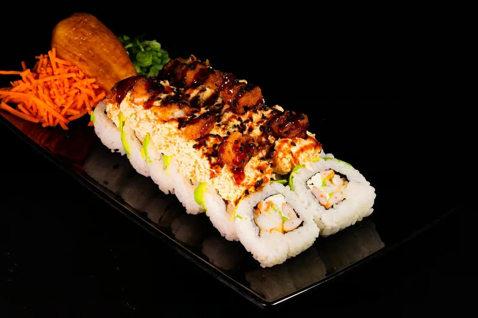 Sushi rolls with shrimp, topped with eel and sauce, served with shredded carrots, a piece of sweet potato, and green onions on a black plate.