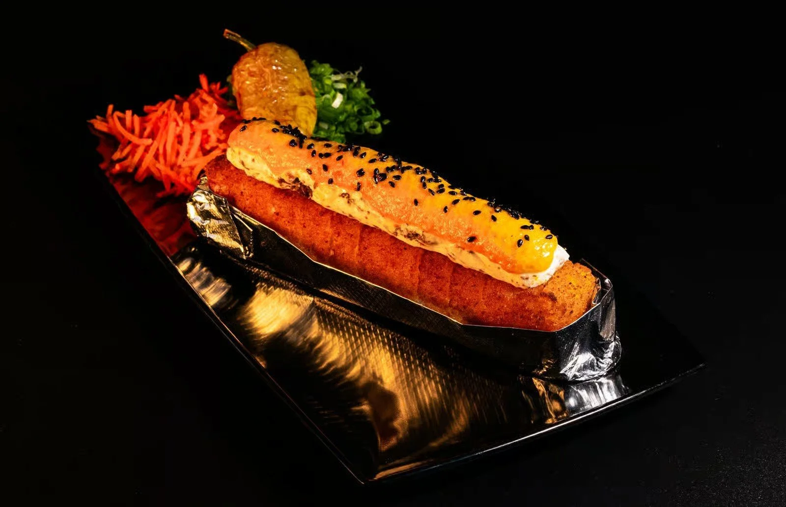 A fish fillet topped with seasoned and black sesame seeds, served with a side of shredded carrots, a small serving of wasabi, and chopped green onions on a black rectangular plate.