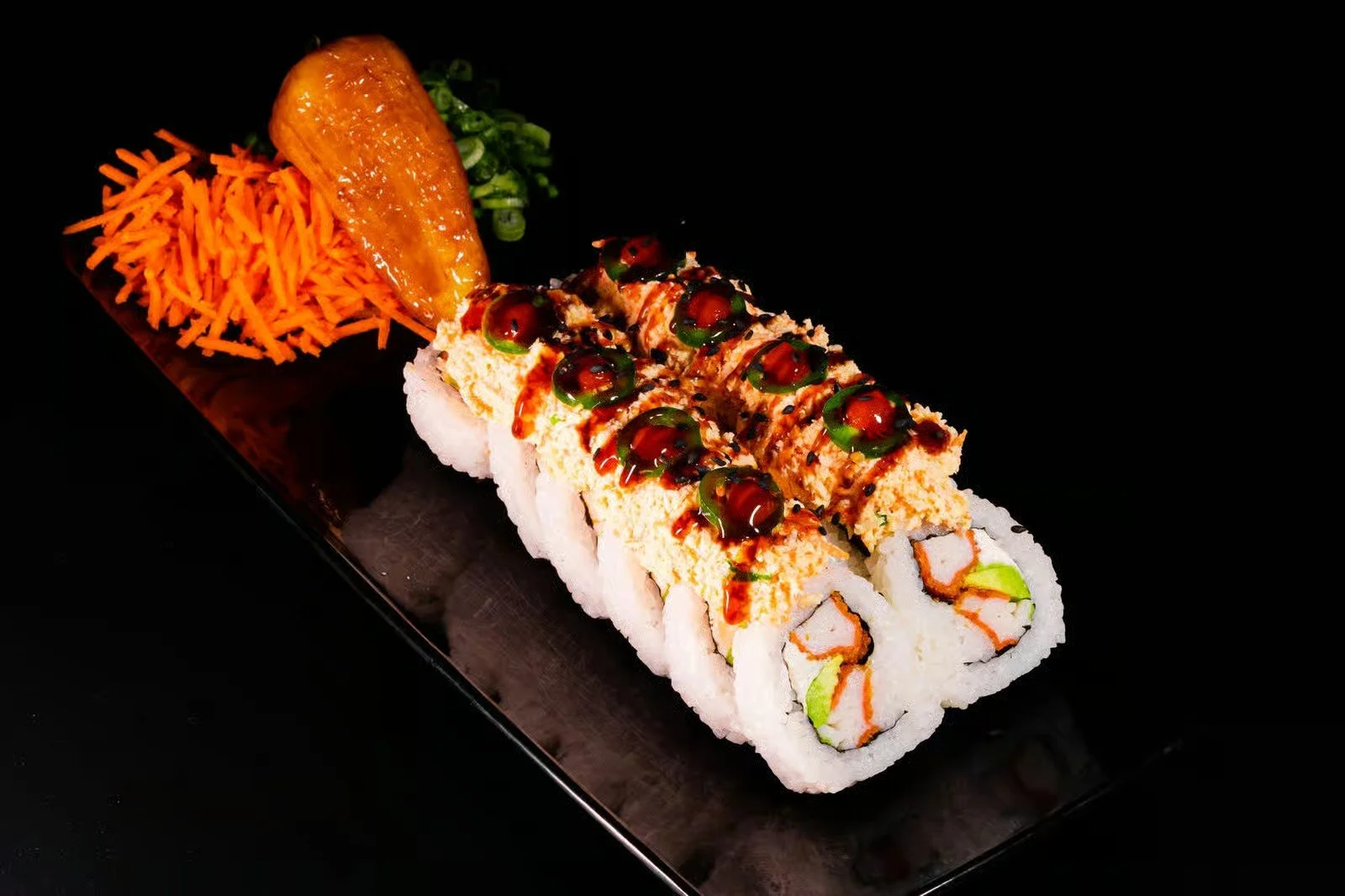 Sushi roll with eel, drizzled with sauce and topped with green spheres, served with shredded carrots, a slice of banana, and chopped green onions on a black plate.