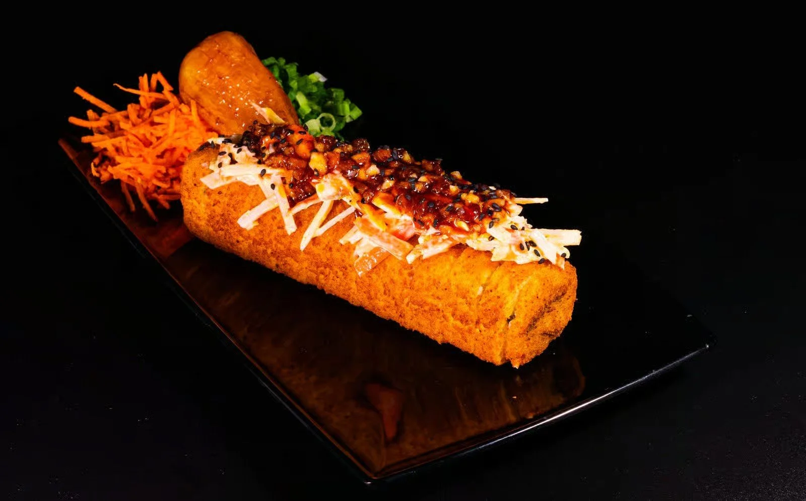 Fried sushi roll topped with shredded cabbage and sauce, served with shredded carrots, green onions, and a fried item on a black rectangular plate.