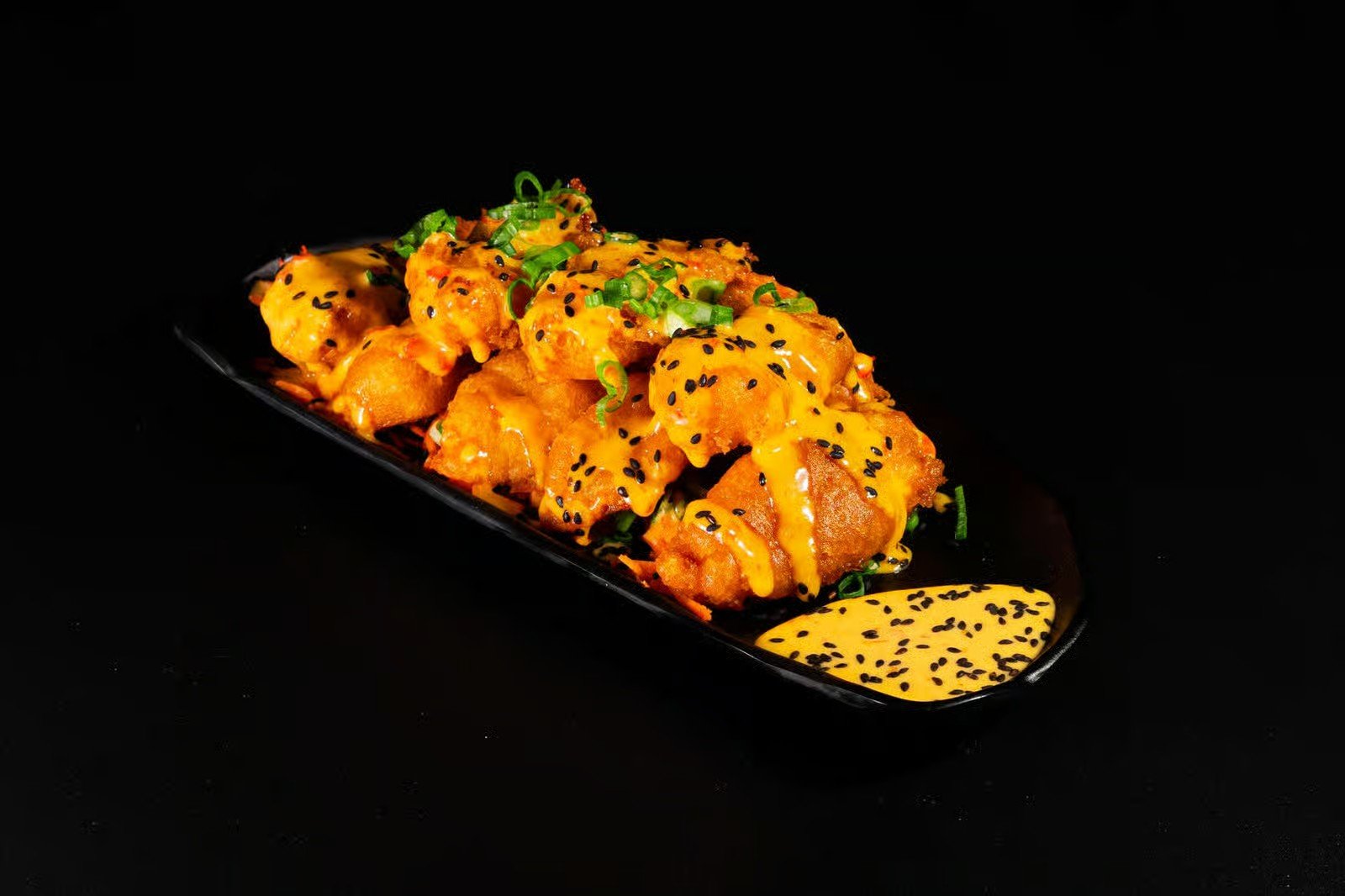 Fried spicy chicken pieces topped with chopped green onions and black sesame seeds, served with a yellow dipping sauce with black sesame seeds, on a black rectangular plate against a black background.