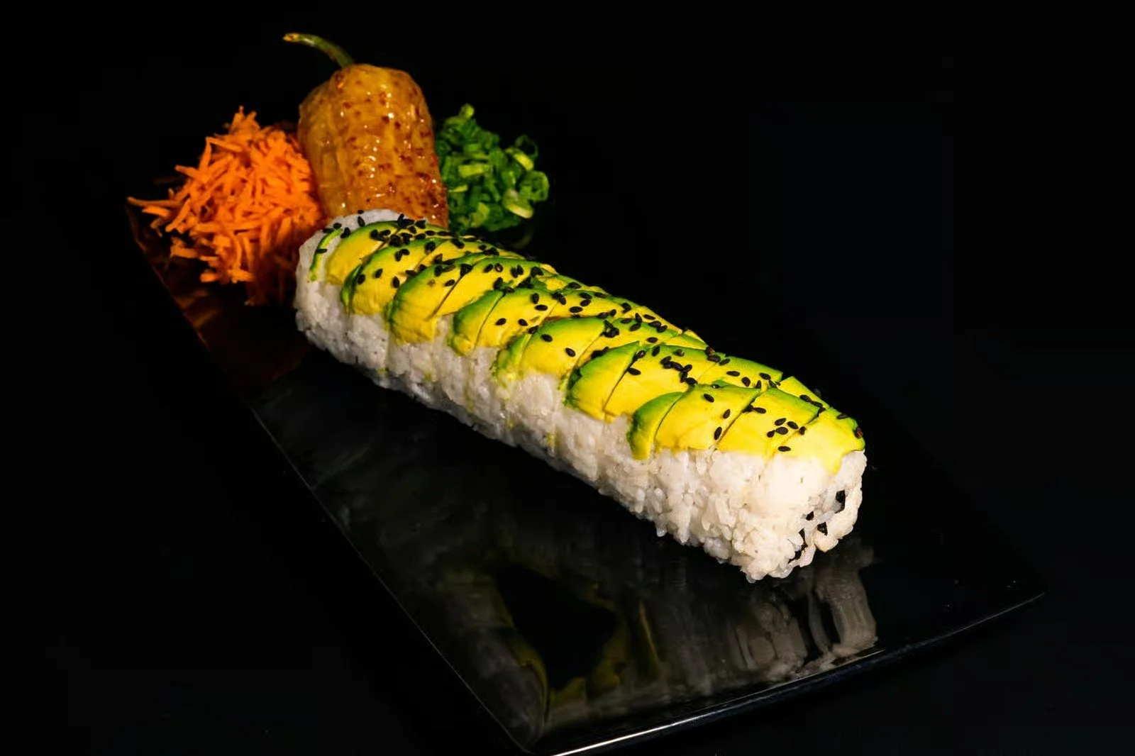Sushi roll topped with sliced avocado and black sesame seeds, accompanied by shredded carrots, a roasted jalapeño, and chopped green onions on a black plate.