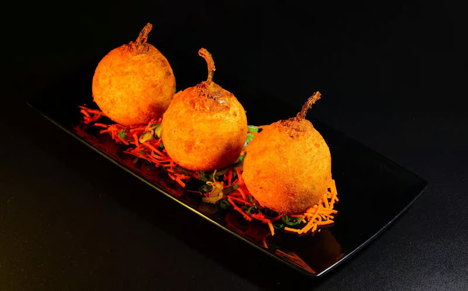 Three deep-fried ball-shaped food items with stems, arranged on a black rectangular plate with shredded orange carrots and green herbs underneath, against a black background.