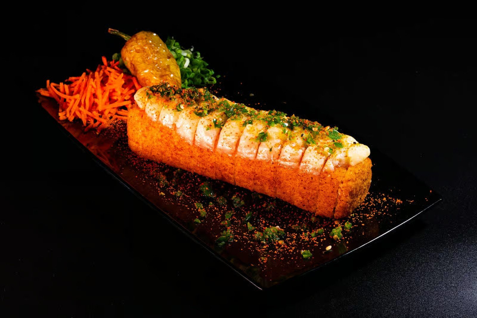 Deep-fried sushi roll topped with sliced fish, green onions, and sauces, served with shredded carrots, chopped green onions, and a roasted yellow pepper on a black rectangular plate.
