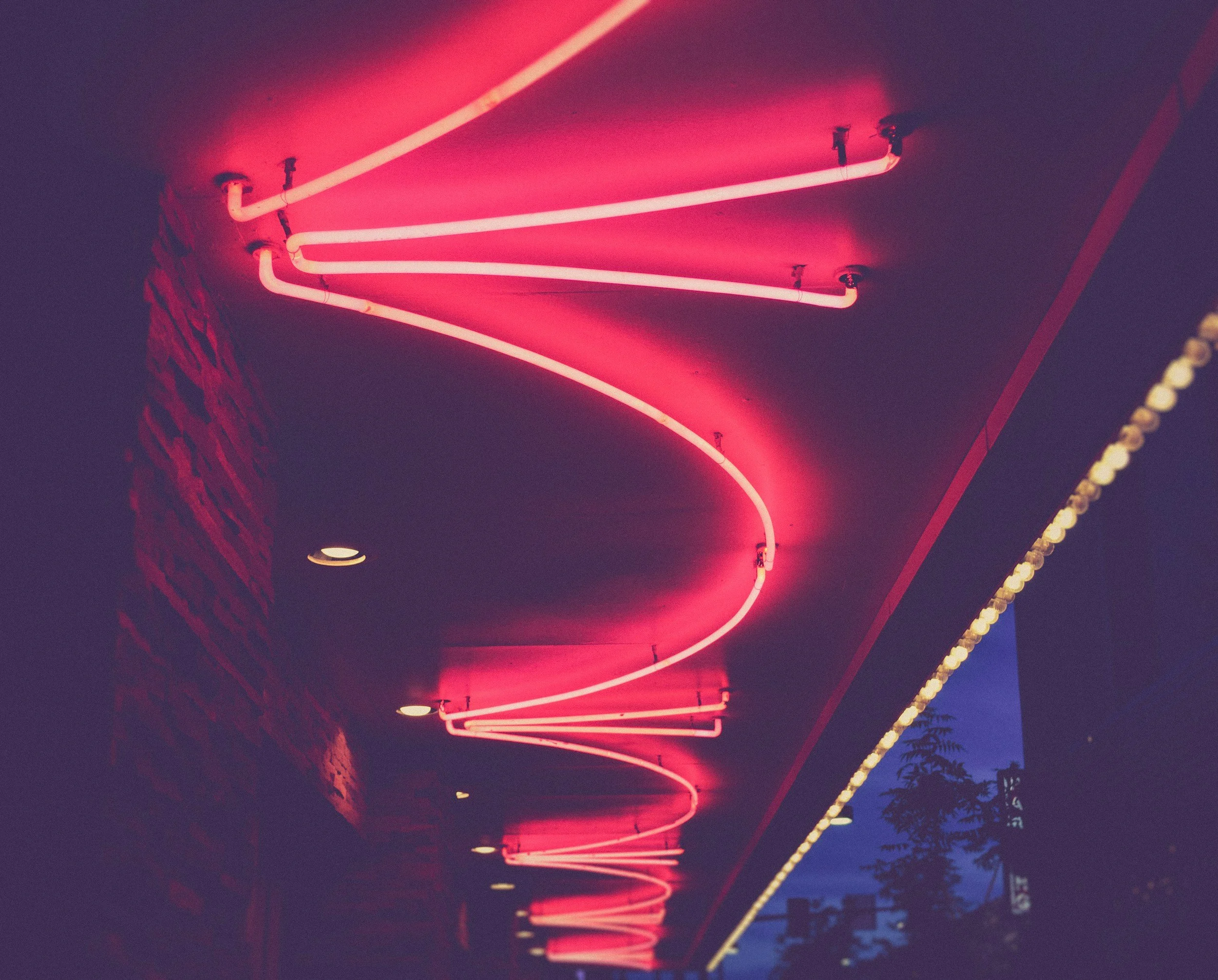 Pink neon light wave sign on a dark exterior wall with trees and a blue evening sky in the background.