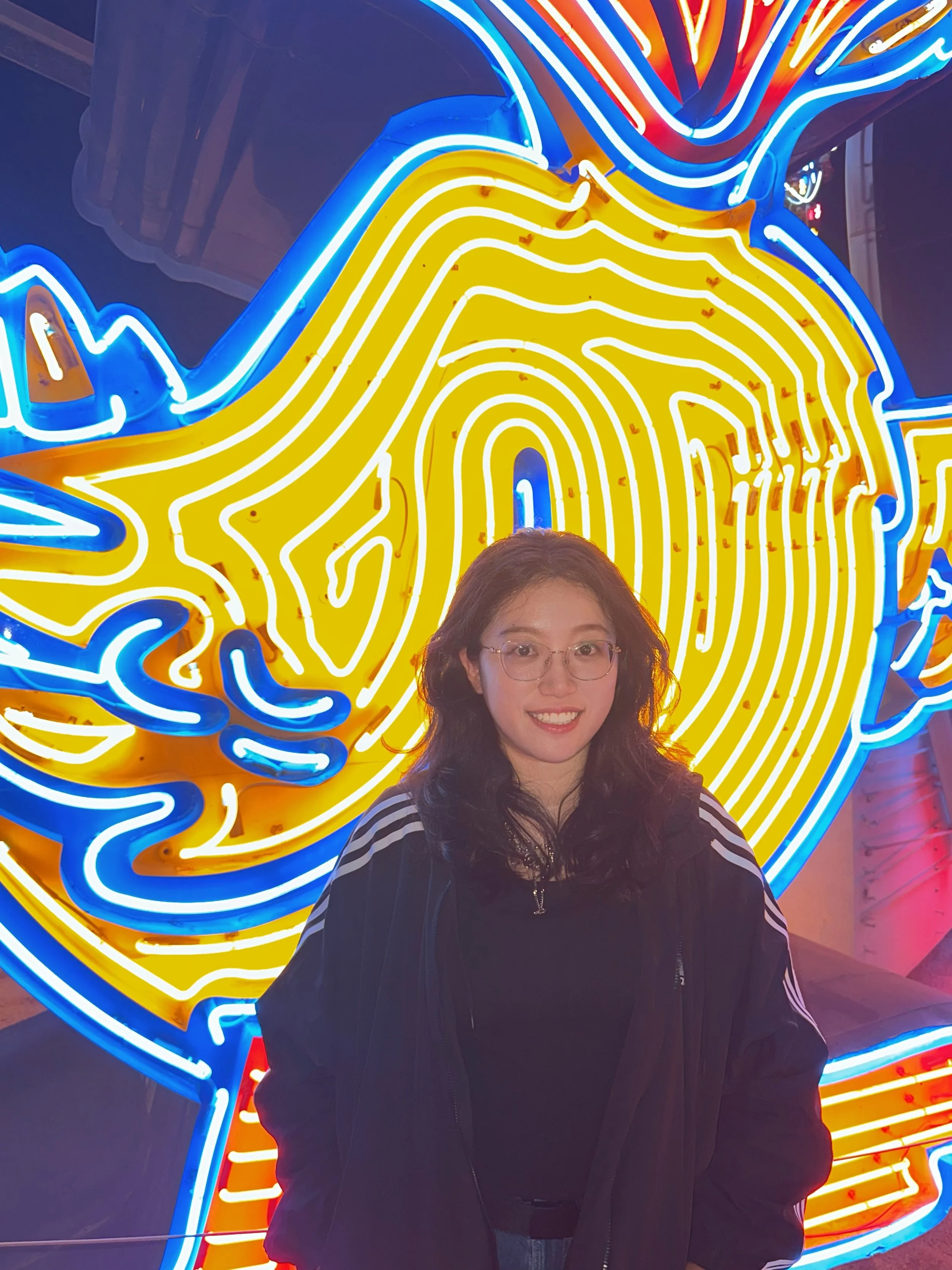 A woman with curly brown hair and glasses smiling at the camera, standing in front of a bright neon sign shaped like a banana.