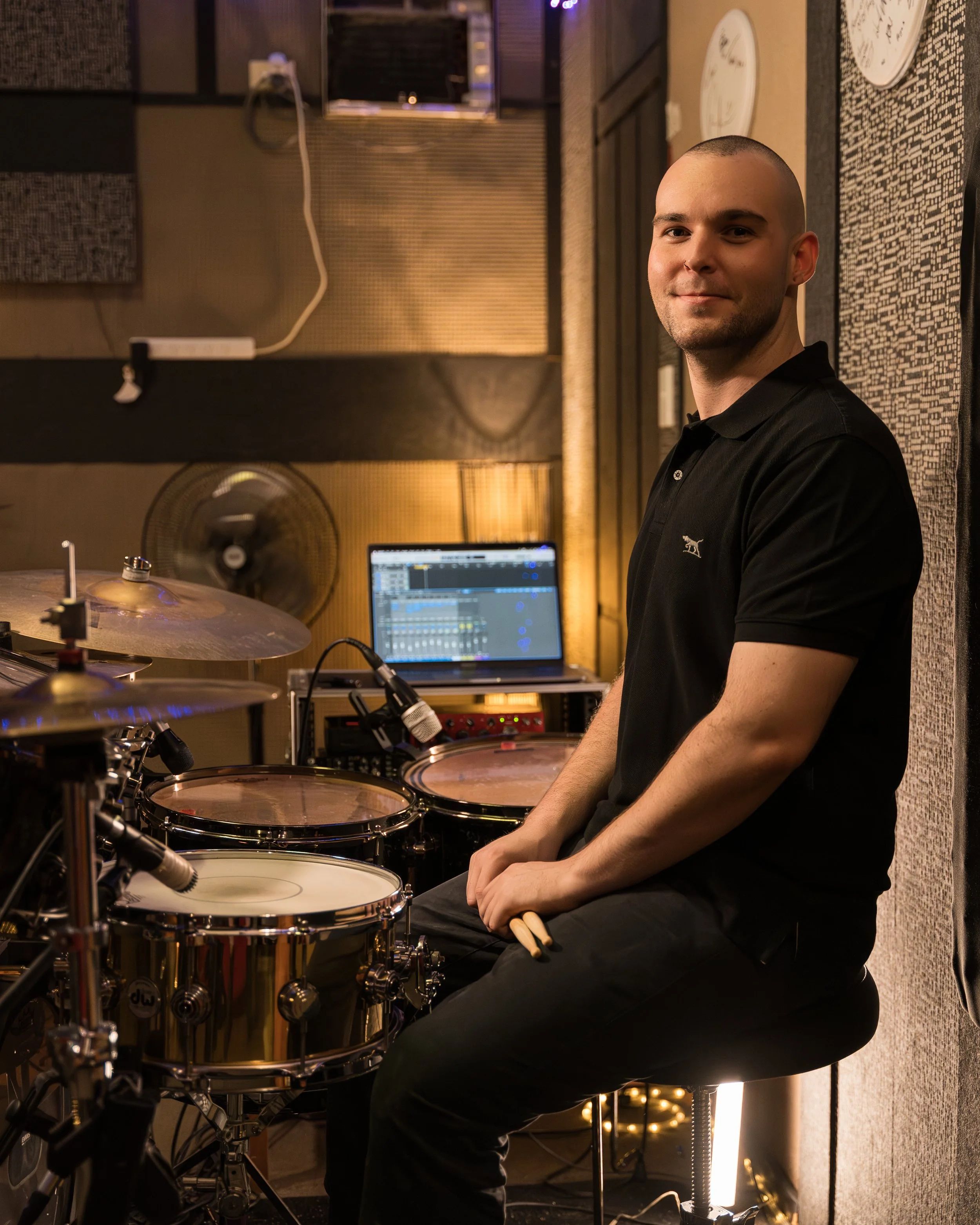 A man sitting on a stool in a music studio with electronic drum pads, a laptop, and a microphone, smiling at the camera.