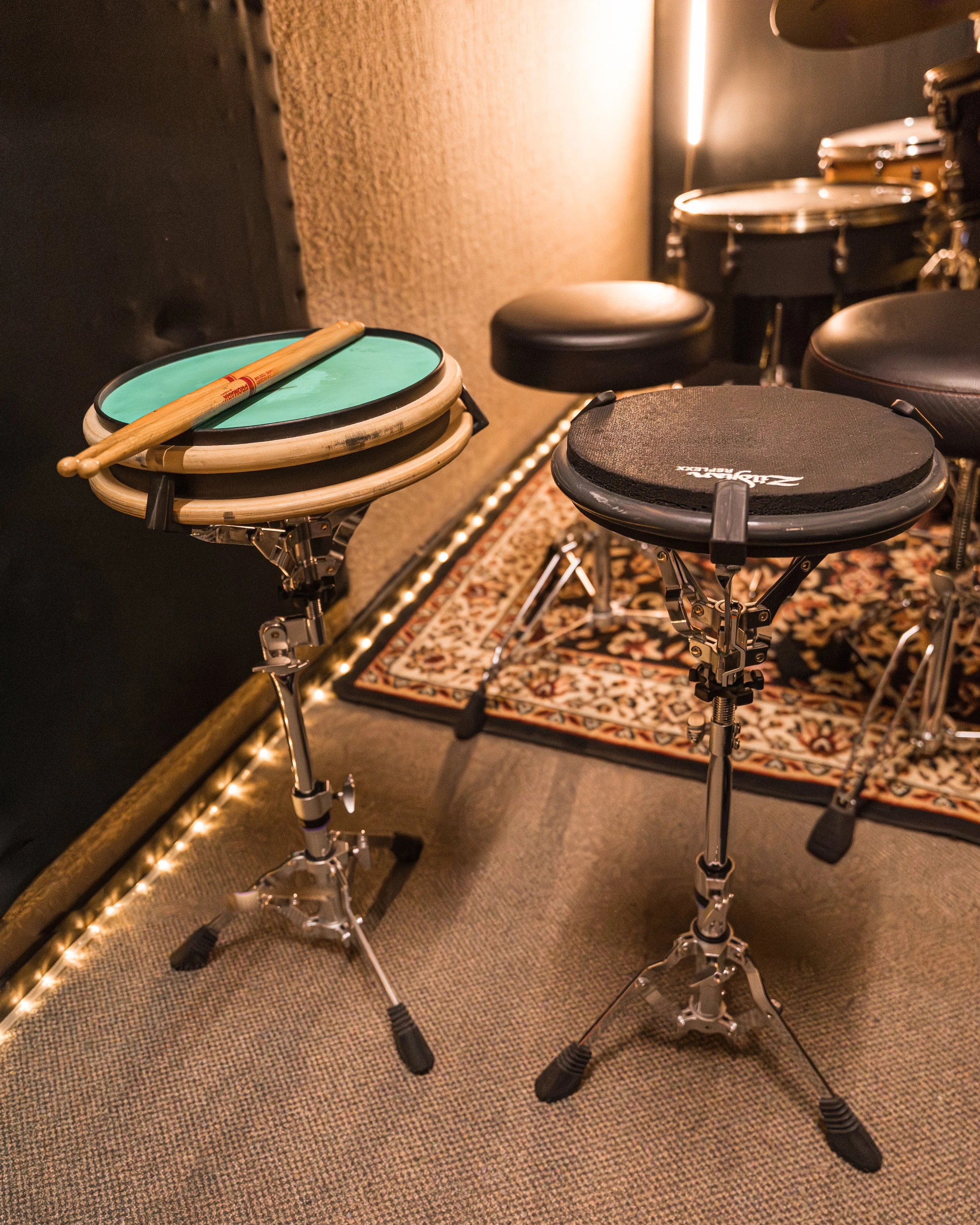 A close-up of a music rehearsal space featuring electronic drum pads, a drumstick resting on a green pad, and other drums in the background, along with a patterned rug on the floor.