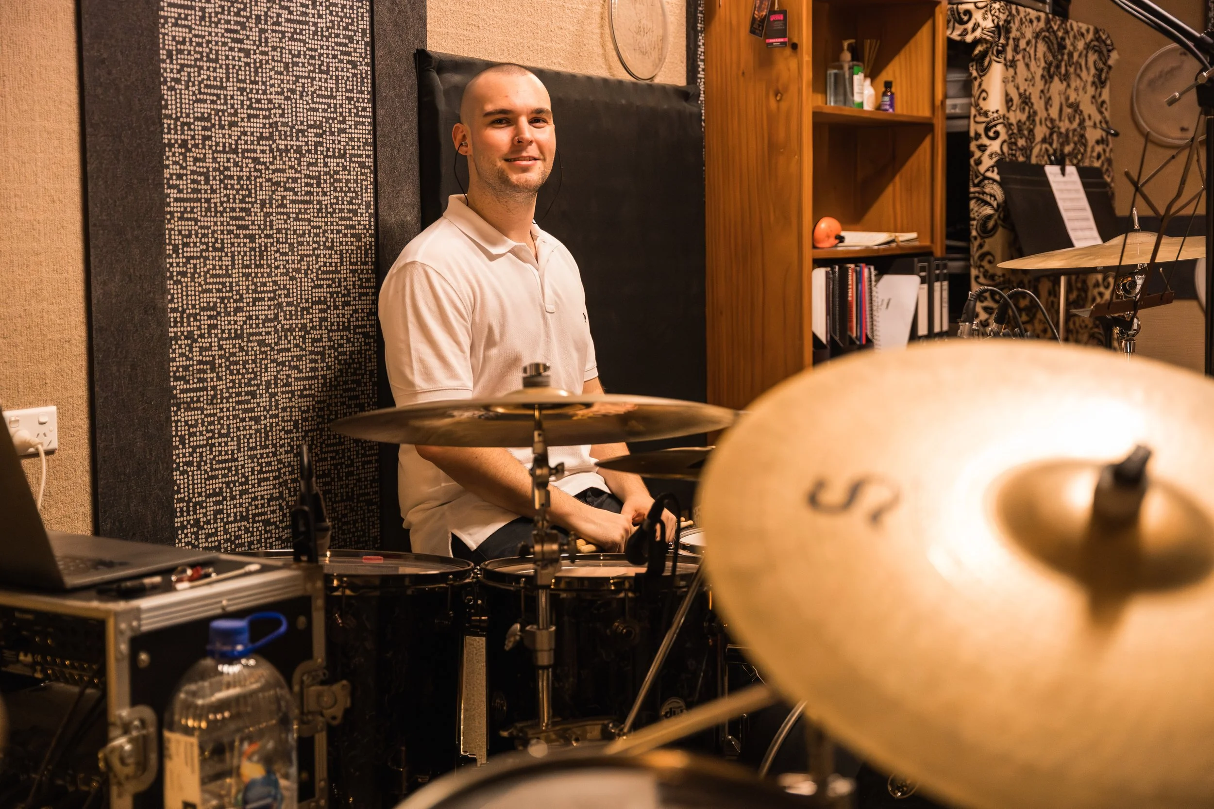 A smiling man with a shaved head wearing a white polo shirt, sitting behind a drum set in a music studio, with musical equipment and shelves in the background.