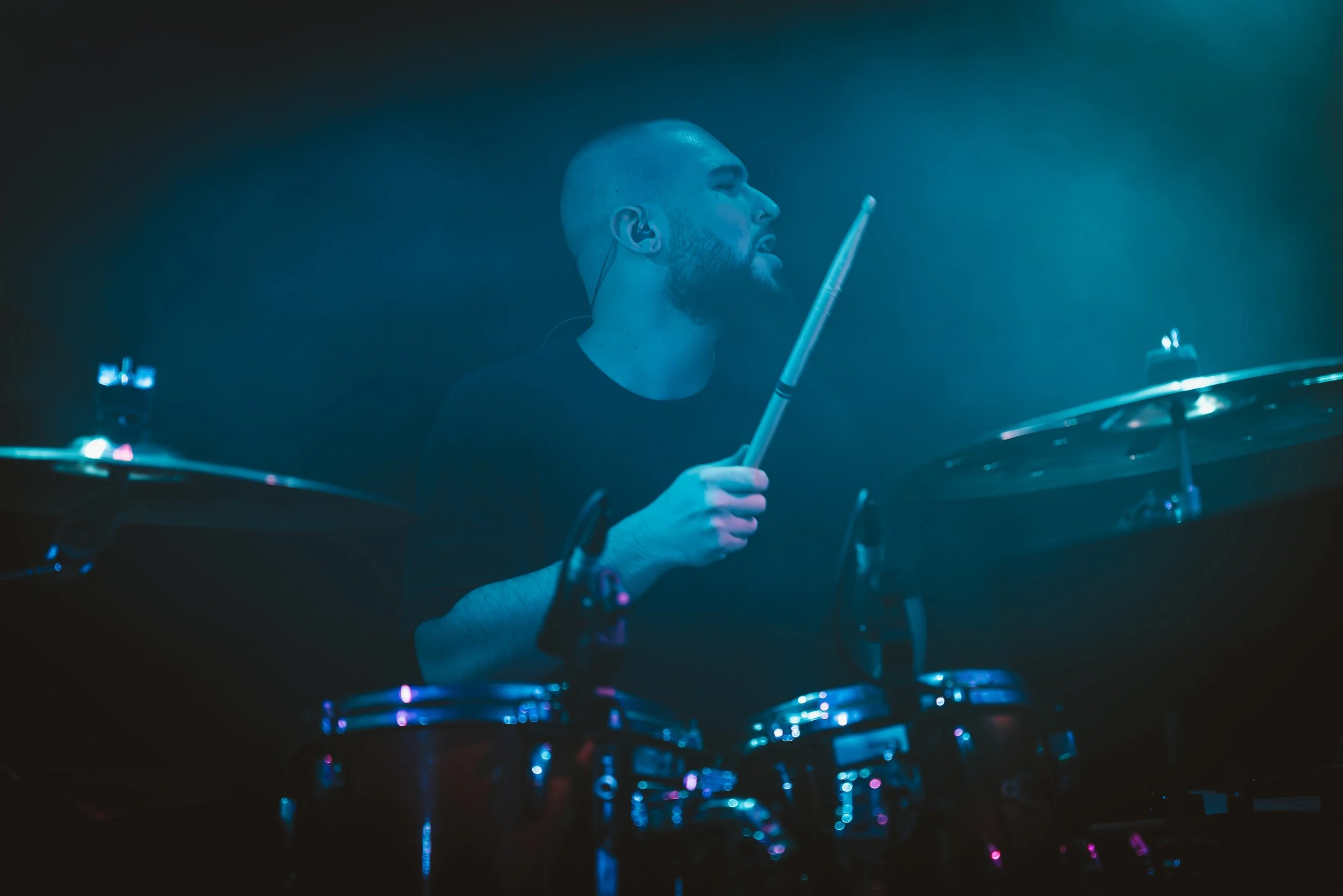 A male drummer with a beard, wearing a black shirt, playing drums with a drumstick in his right hand. He is in a dimly lit setting with blue lighting.