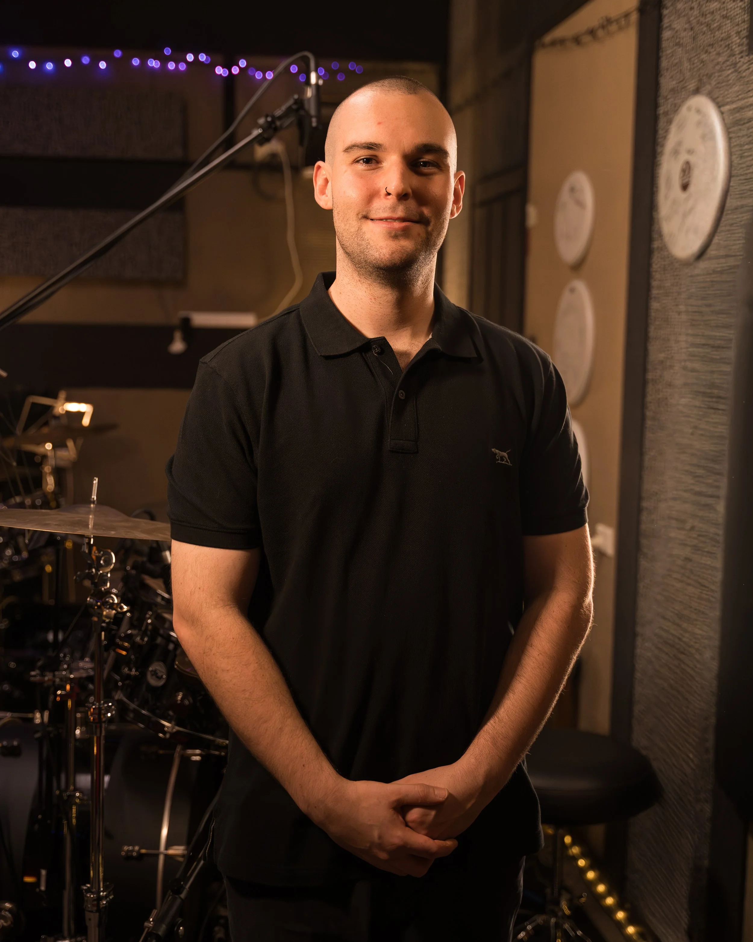 A young man with a shaved head and nose piercing stands in a music studio with drums and music equipment in the background.