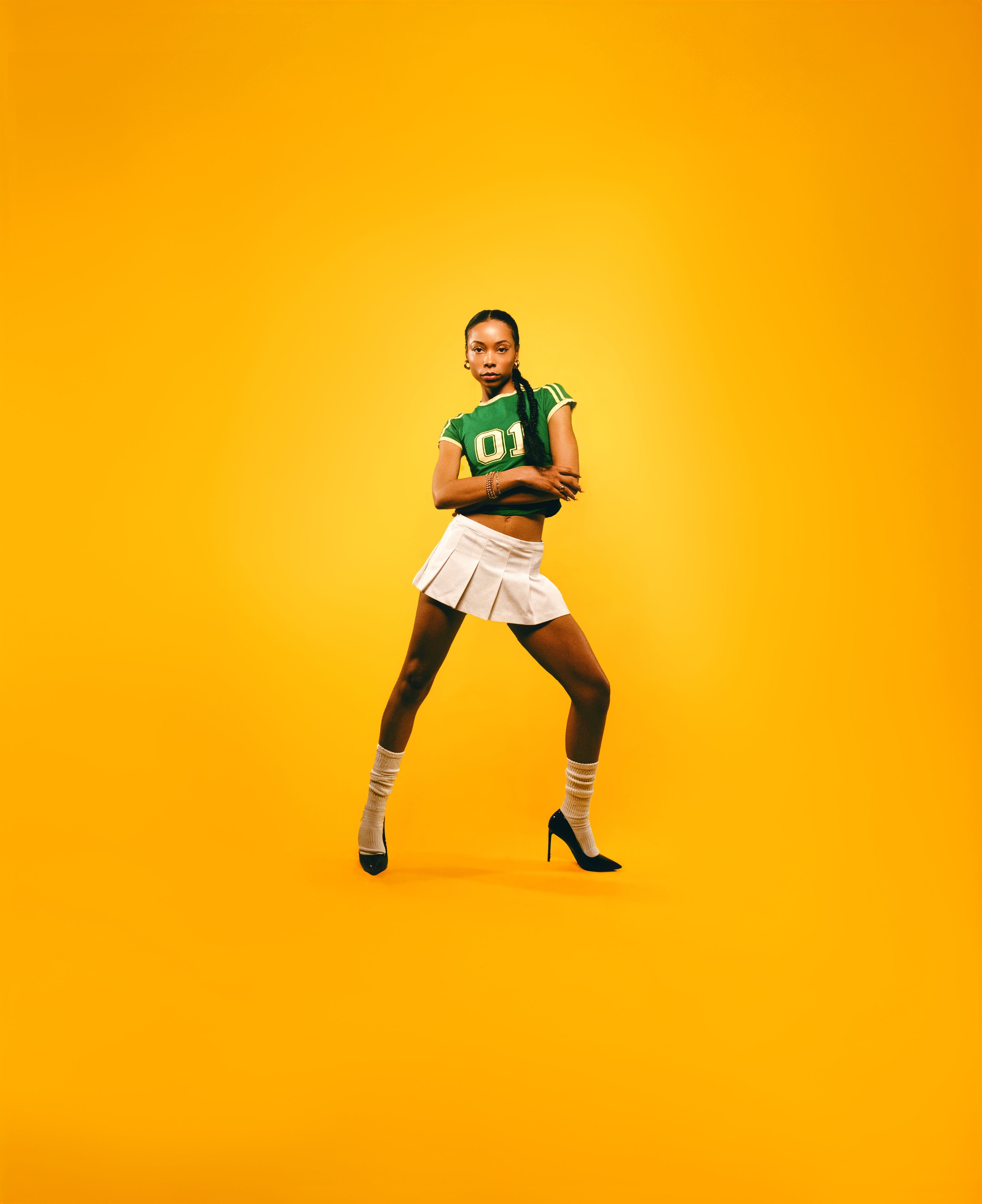 A young woman in a green athletic crop top, beige pleated skirt, and high heels poses against a yellow background.