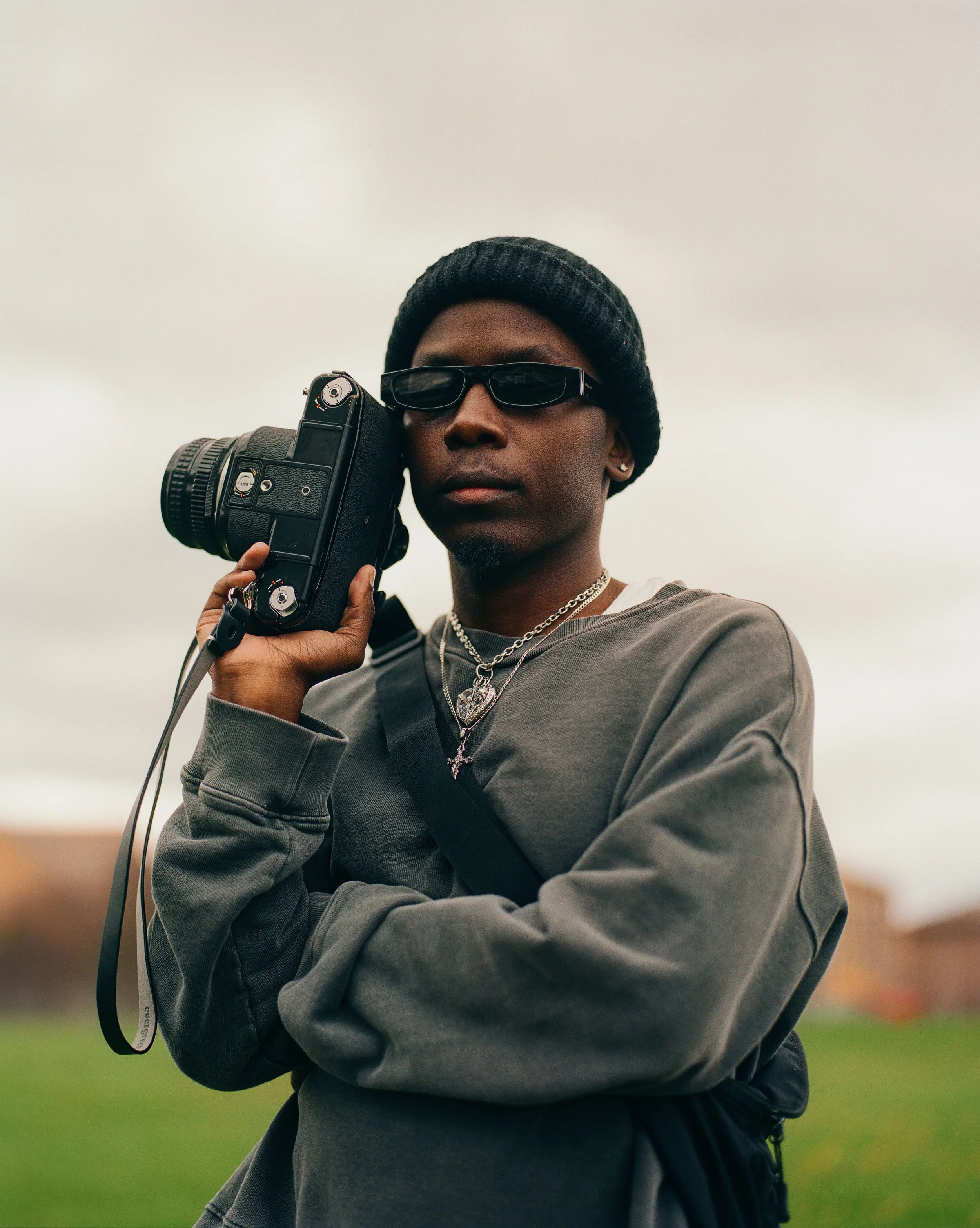 Young man wearing a black beanie and sunglasses holding a camera on his shoulder outdoors.